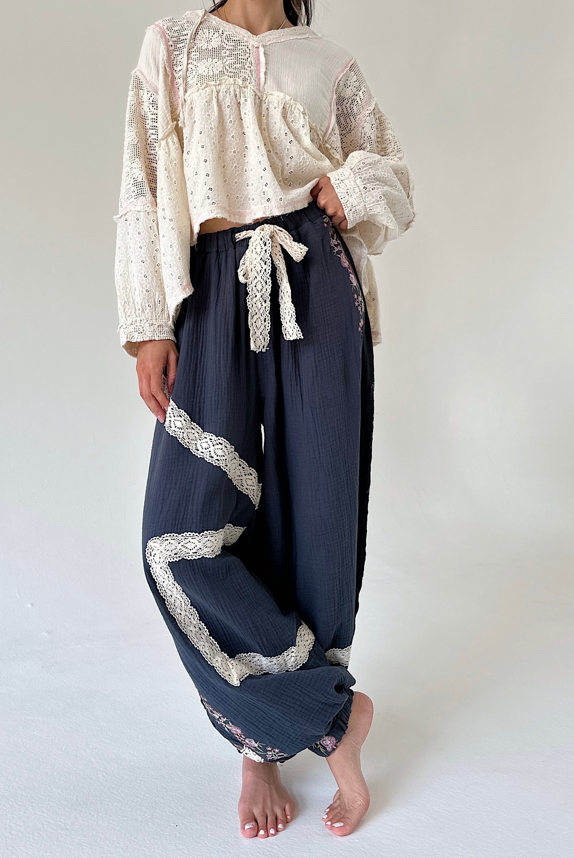 A model wearing a cream-colored lace long-sleeve crop top and dark navy blue crinkled wide-leg pants with white lace trim and a matching tie, standing barefoot against a plain light background.