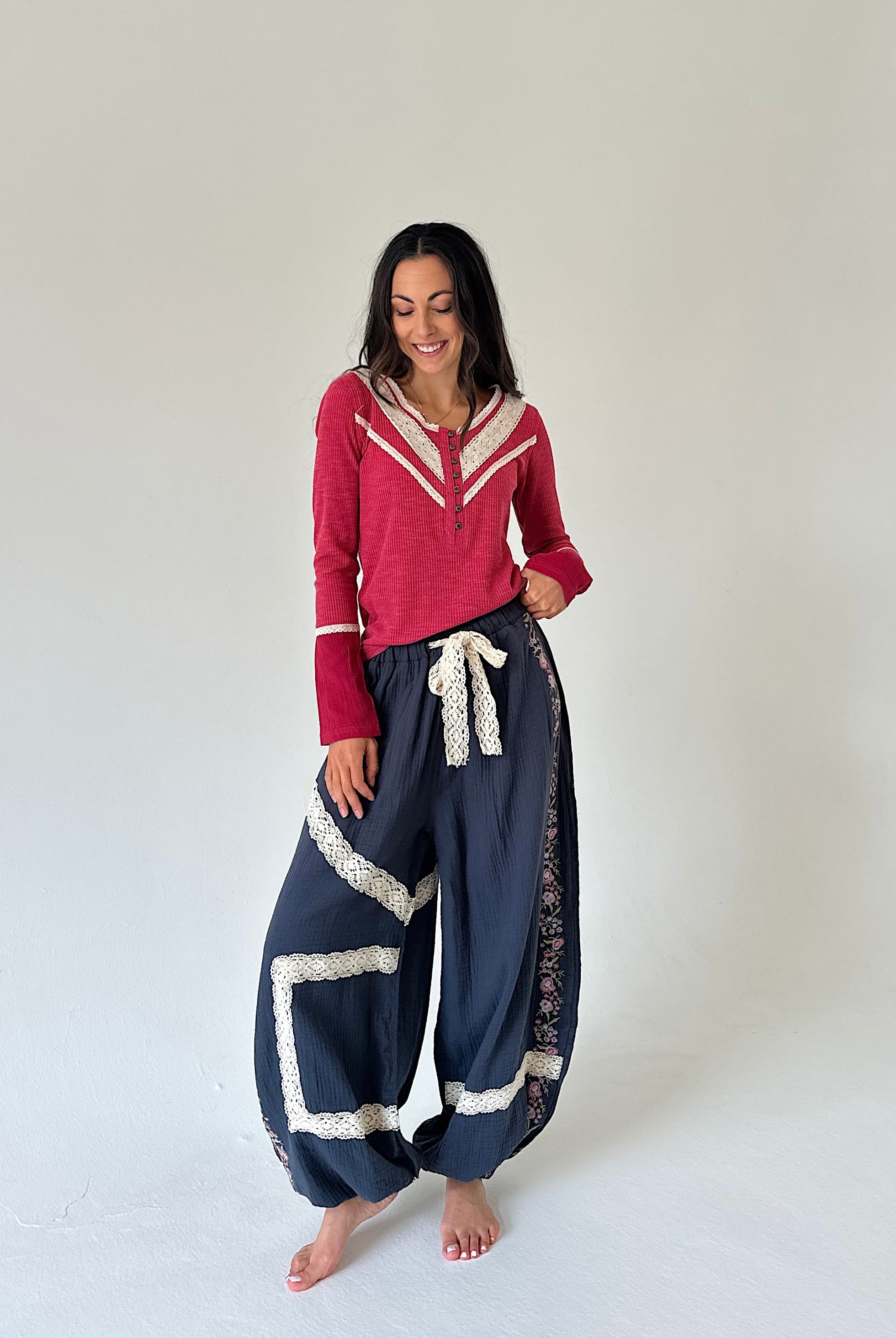 A woman modeling a red long-sleeve top with cream trim and navy blue wide-leg harem pants with cream lace trim, standing against a plain light gray studio background.