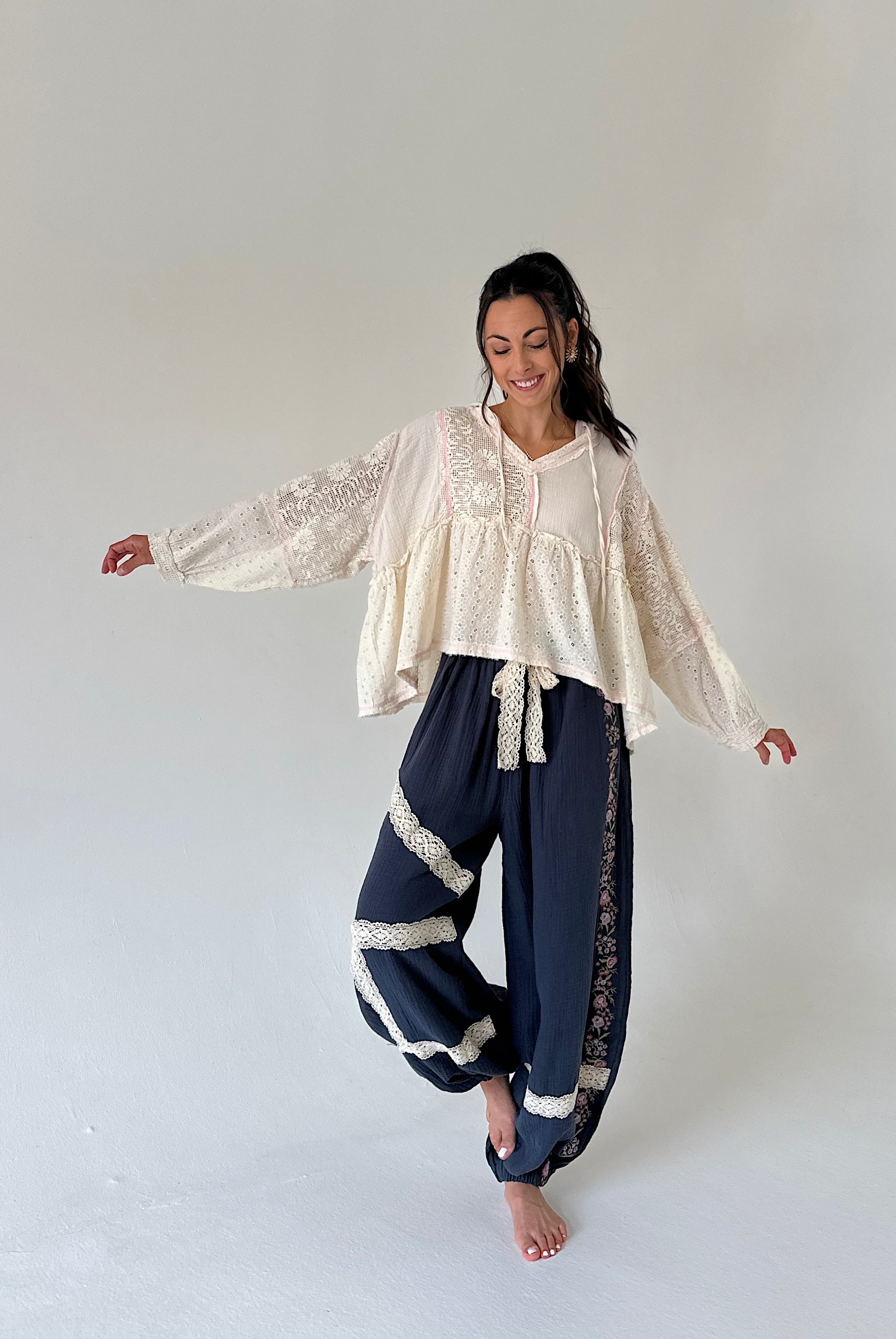 A woman modeling a bohemian-style outfit consisting of a cream lace long-sleeve blouse and navy blue harem pants with cream lace trim, standing against a neutral light gray studio background.