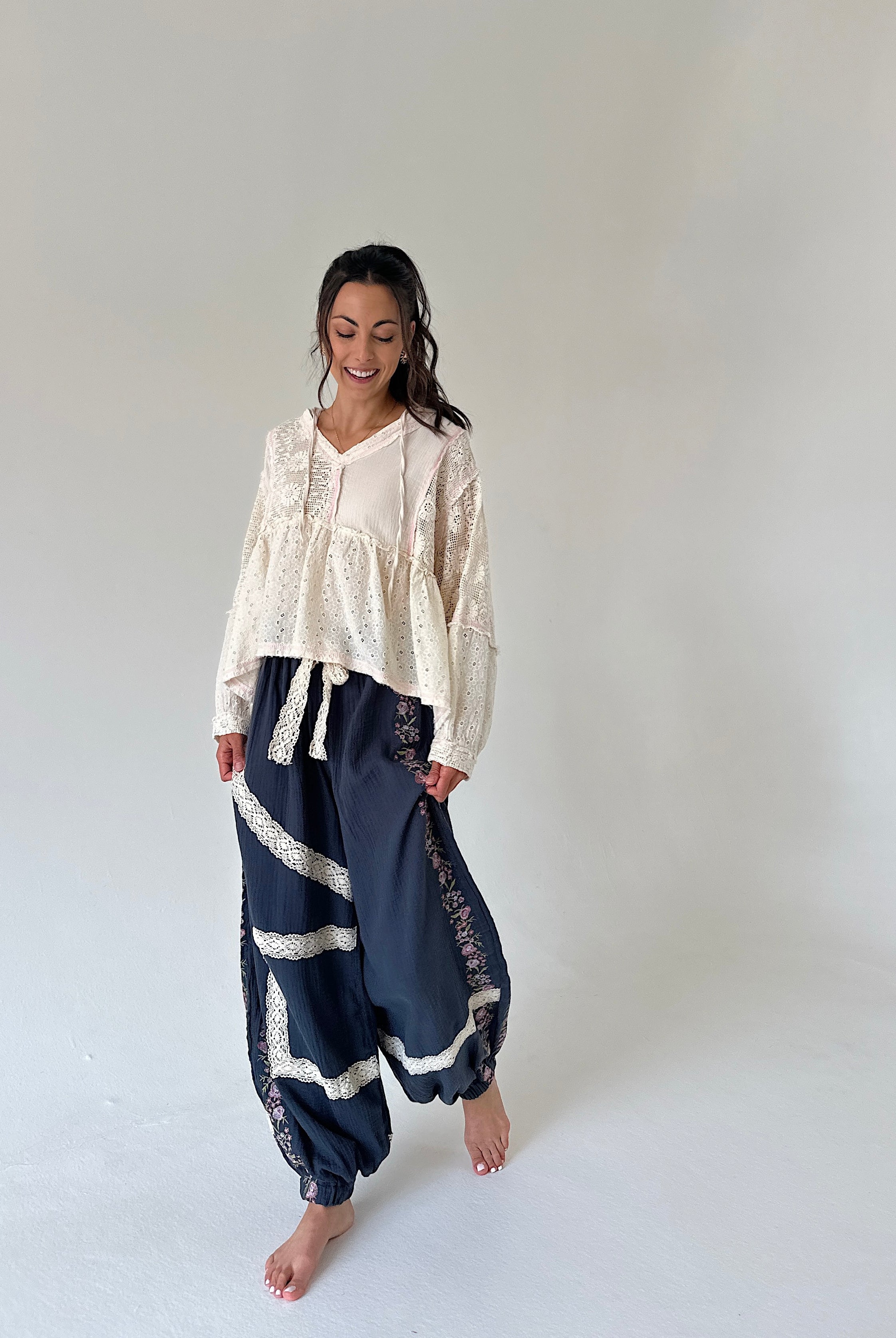 A woman modeling a bohemian-style outfit consisting of a cream lace long-sleeve top and navy blue patterned harem pants with lace trim, standing against a plain light gray studio background.