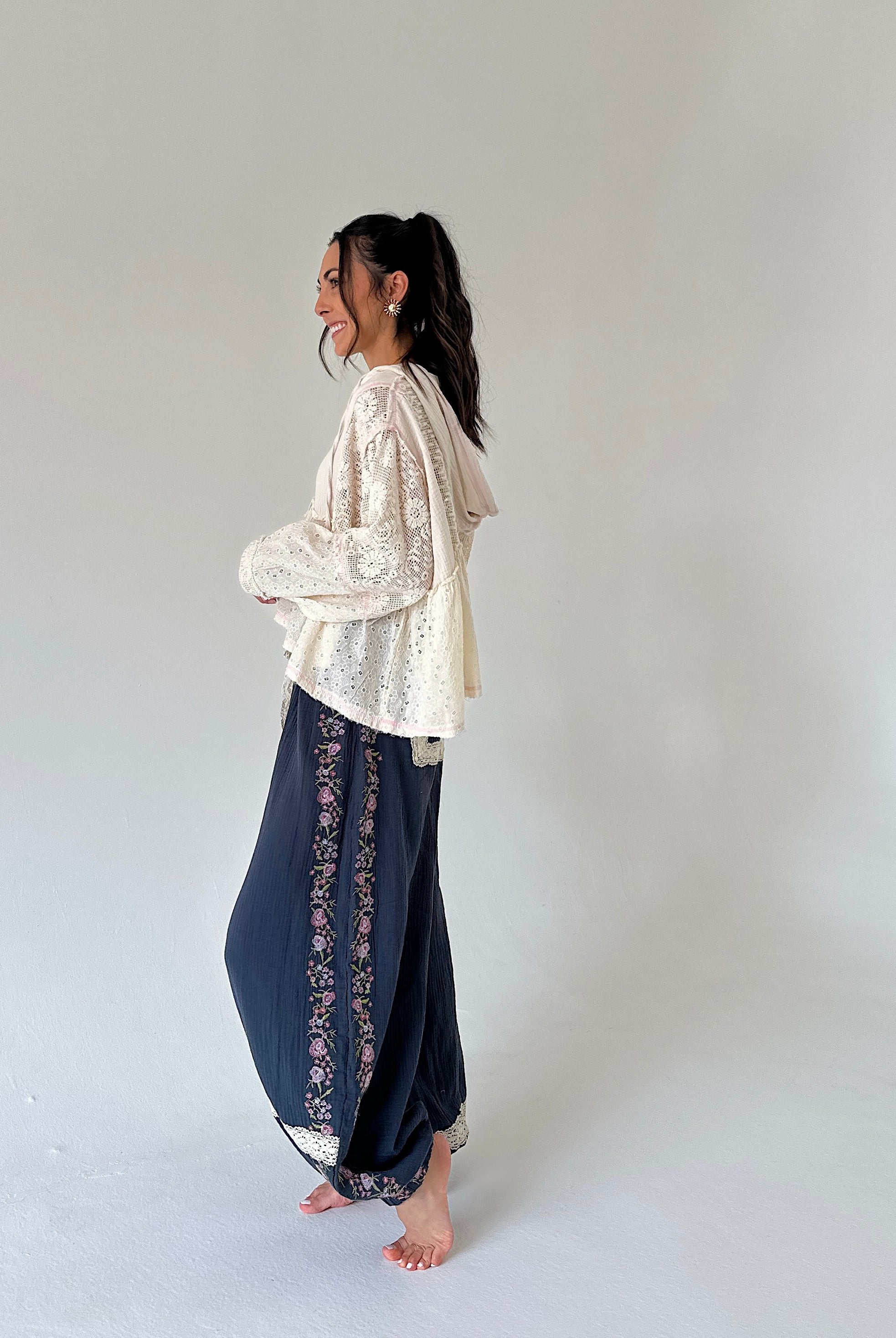 A woman stands barefoot in profile wearing a cream lace long-sleeve top and dark navy wide-leg pants with floral embroidered side panels, against a plain light gray studio background.