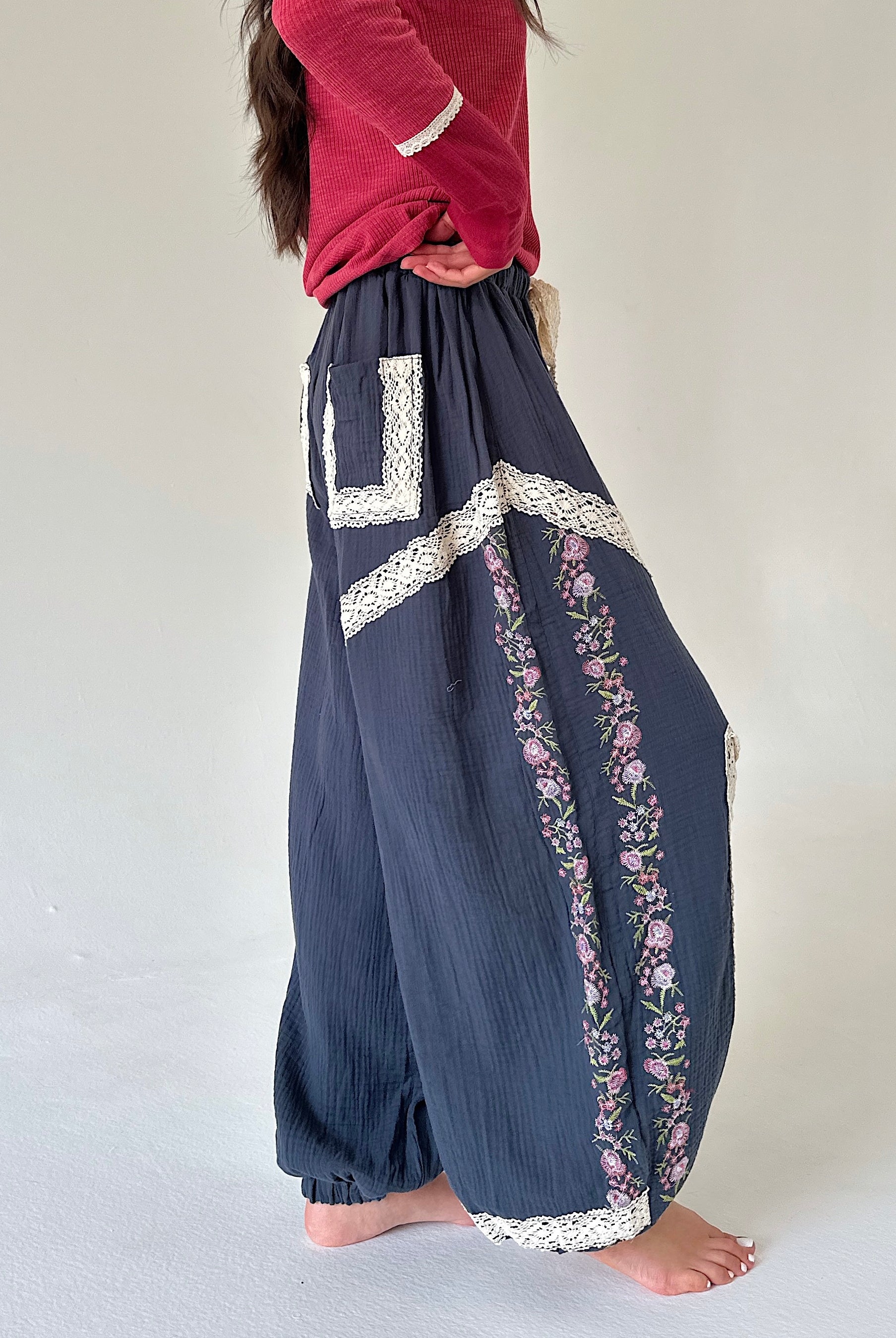 A person wearing loose navy blue harem-style pants with white lace trim and pink floral embroidery, paired with a red top, standing barefoot against a plain light background.