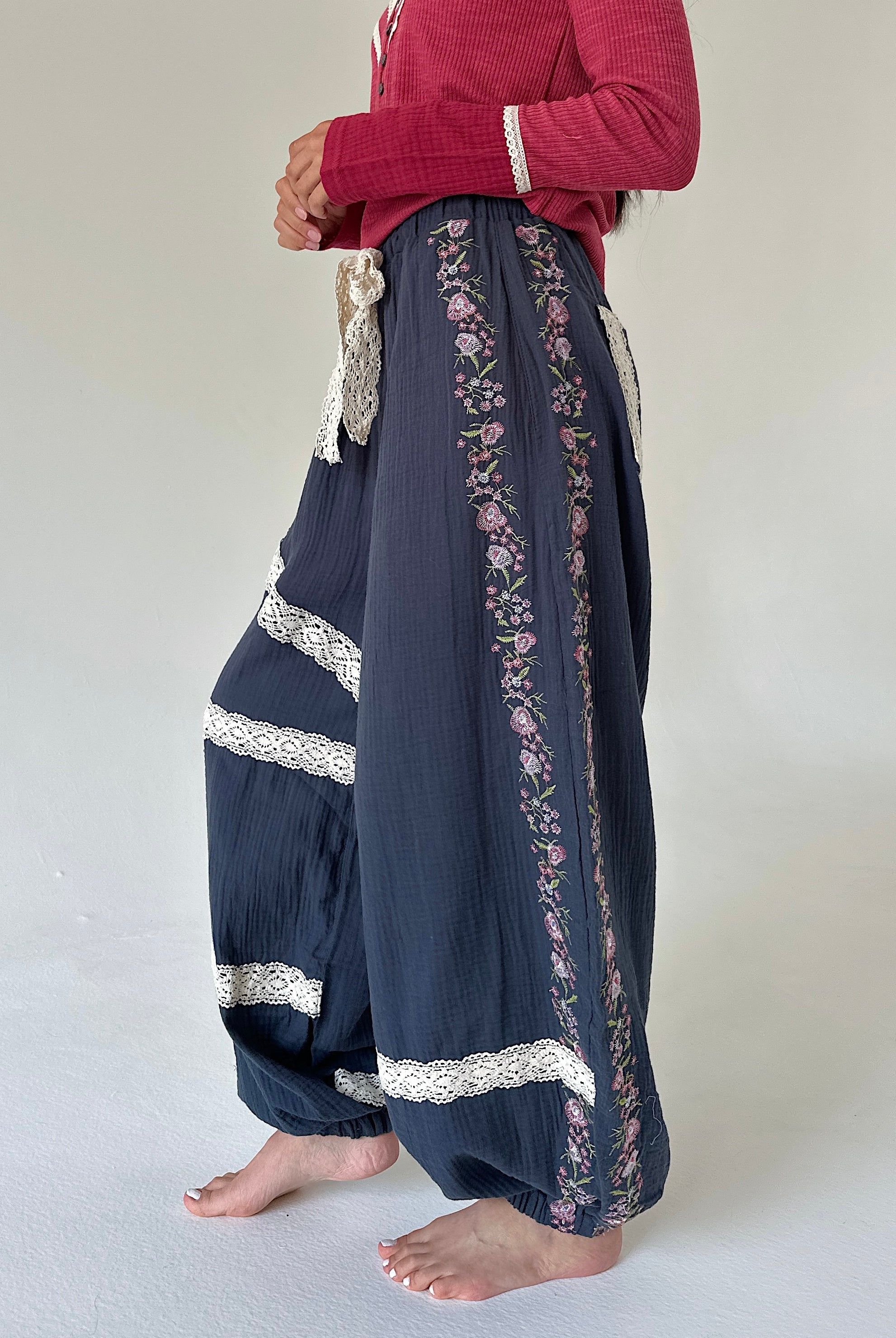 A person wearing loose, wide-leg navy pants with floral embroidery and white lace trim, paired with a red long-sleeve top, standing barefoot against a plain light background.