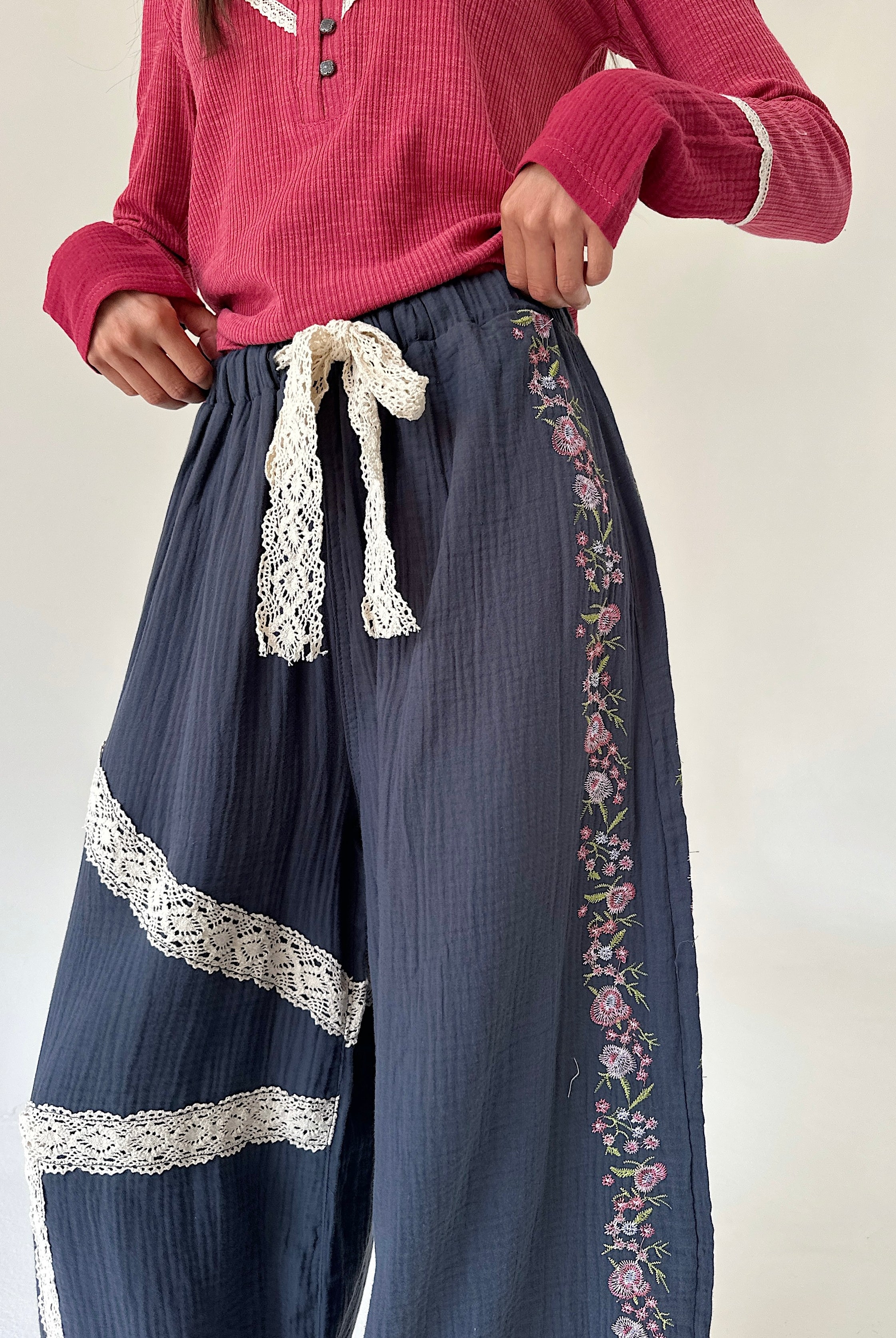 A person wearing loose navy wide-leg pants with cream lace trim, floral embroidery down the side, and a matching cream lace tie at the waist, paired with a pink ribbed top against a plain light background.