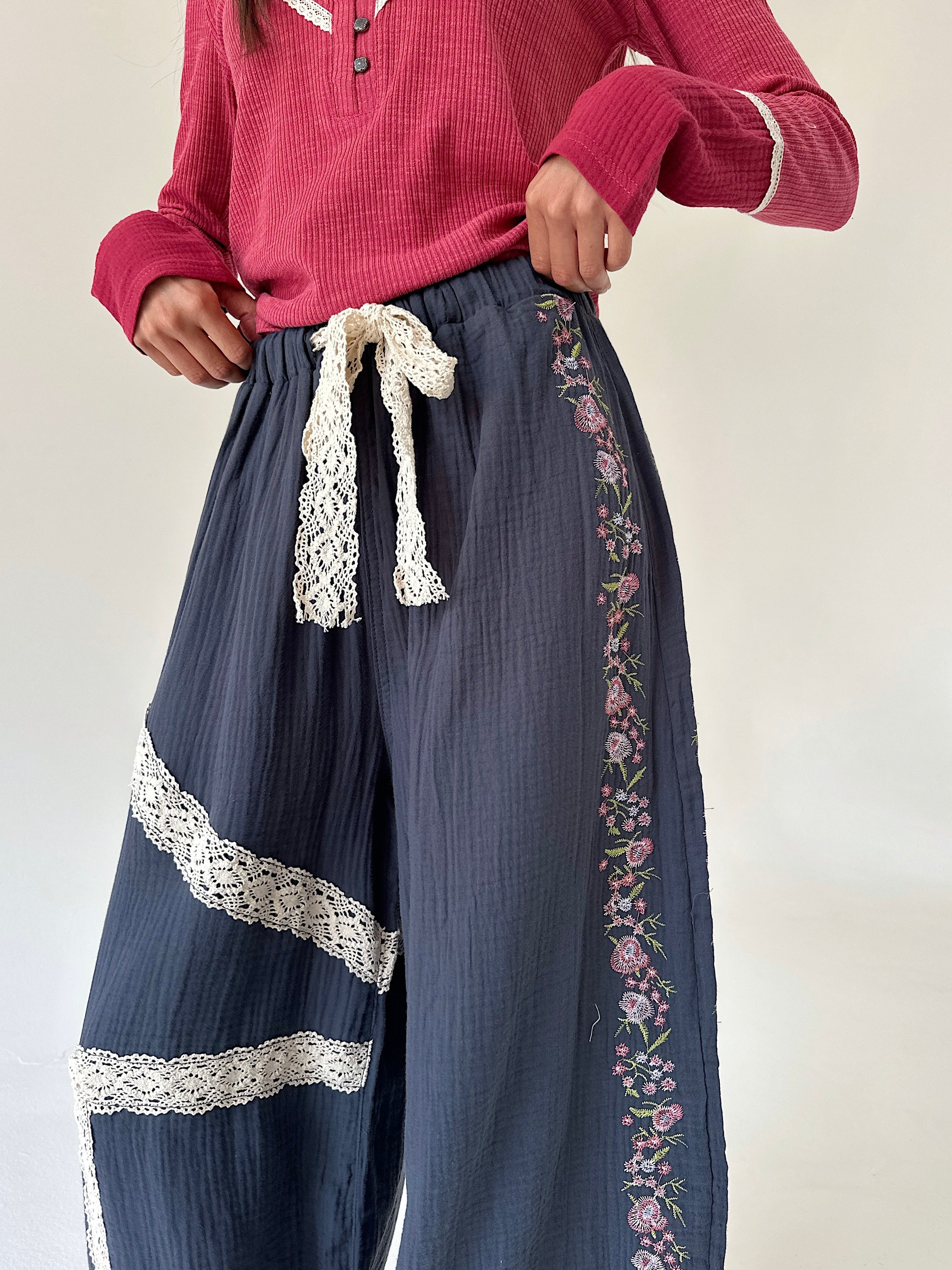 A person wearing loose navy wide-leg pants with cream lace trim, floral embroidery down the side, and a matching cream lace tie at the waist, paired with a pink ribbed top against a plain light background.