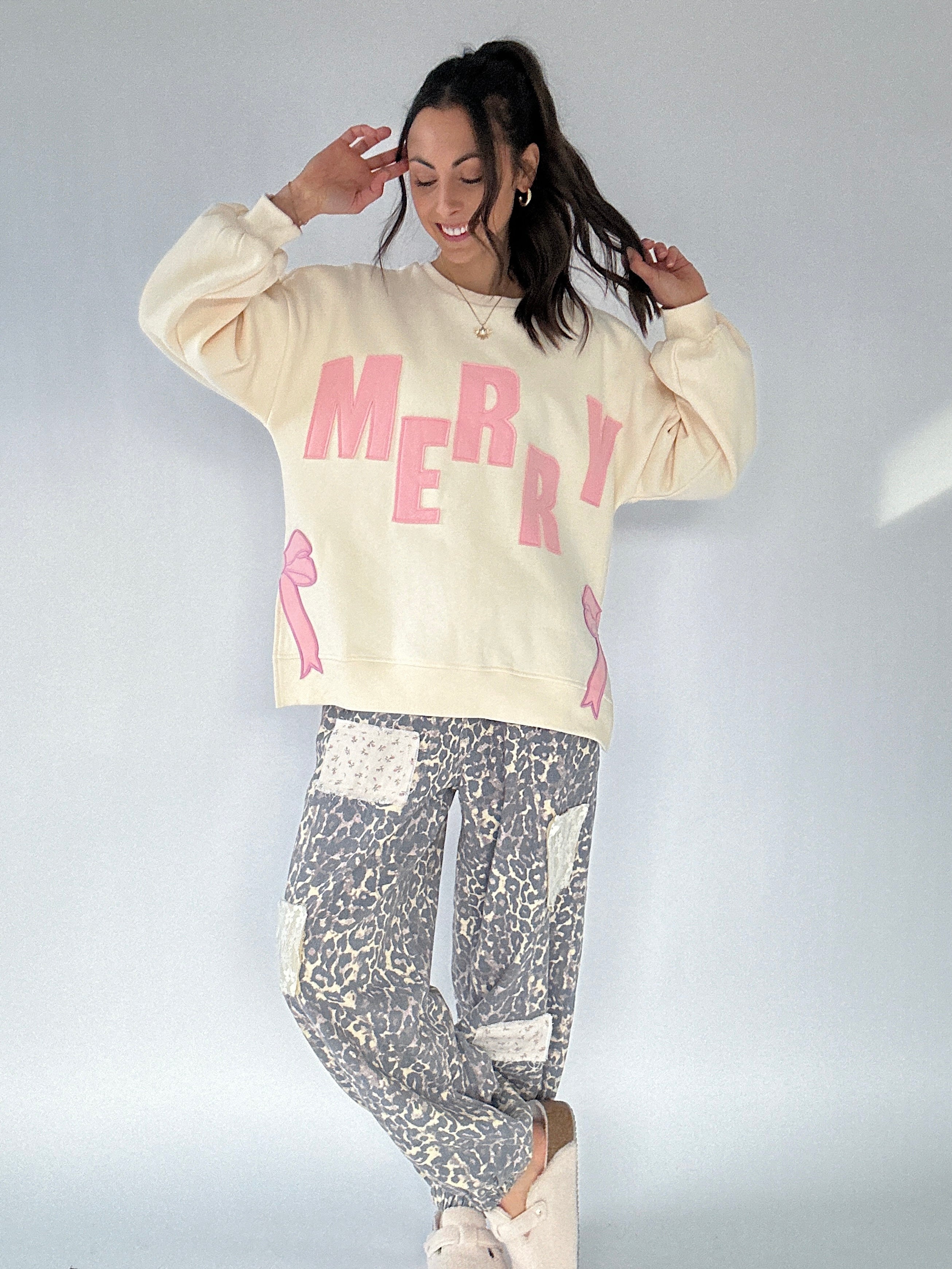 Person wearing a cream sweatshirt with 'MERRY' in pink letters and leopard patterned pants on a plain background