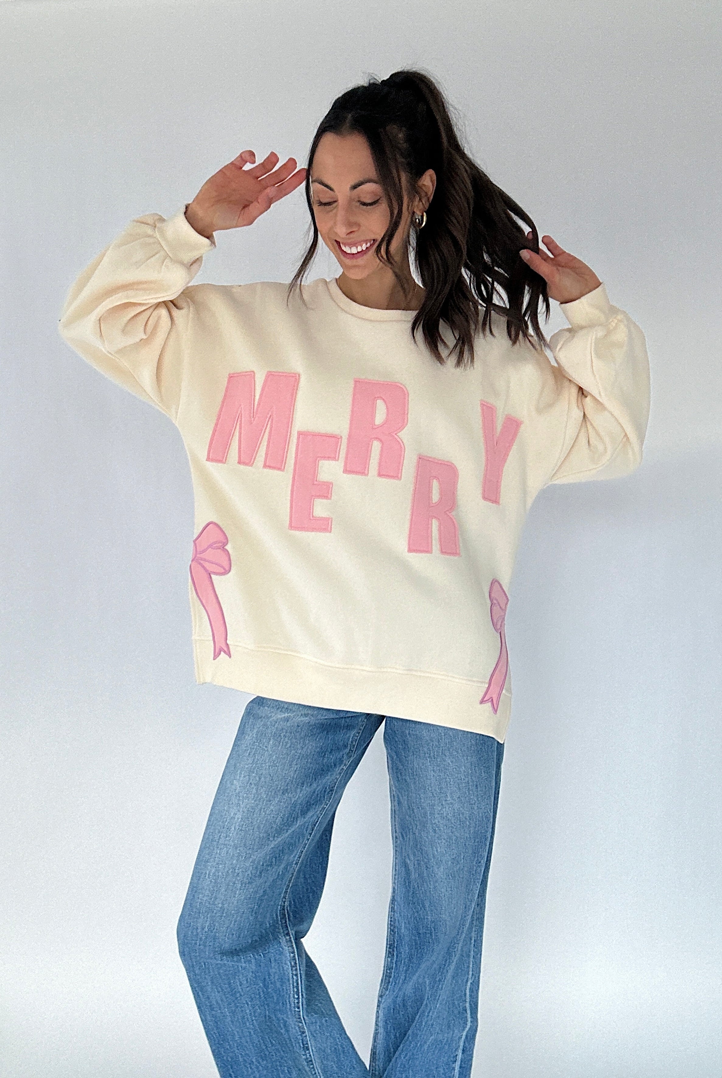 Person wearing a cream sweatshirt with pink "MERRY" text and graphics on a plain background