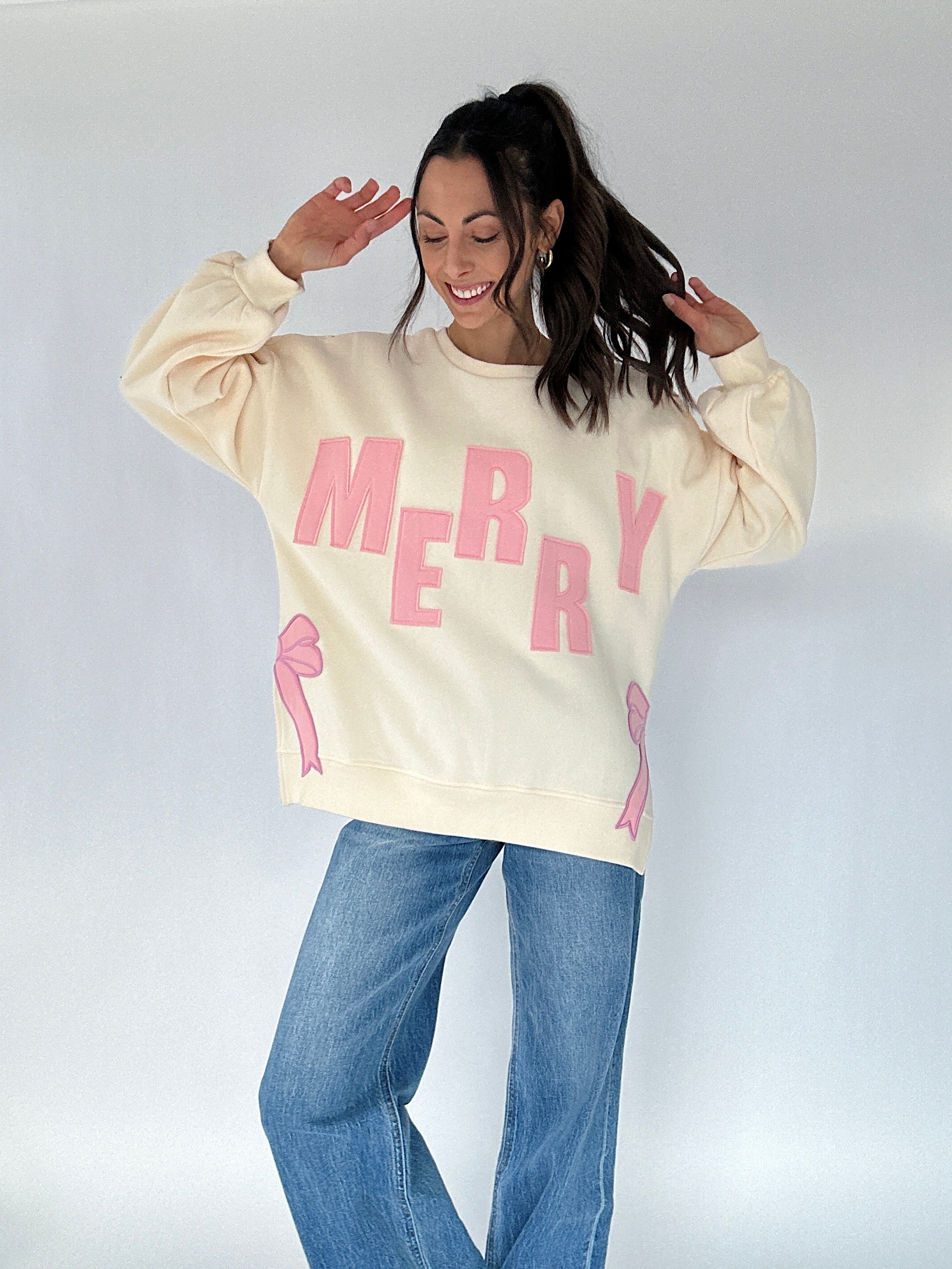 Person wearing a cream sweatshirt with pink "MERRY" text and graphics on a plain background