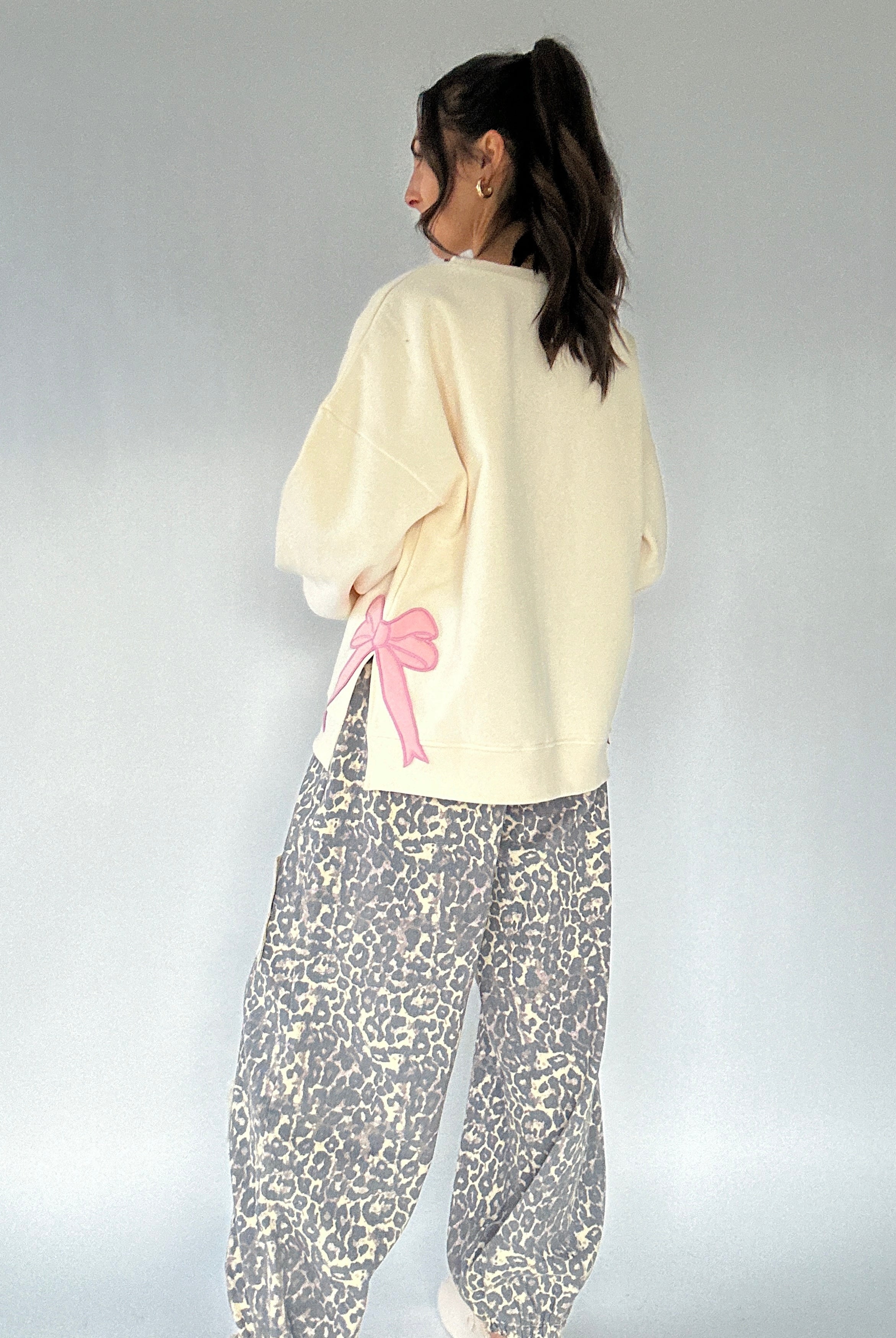 Person wearing a cream sweatshirt with a pink bow and leopard patterned pants on a plain background