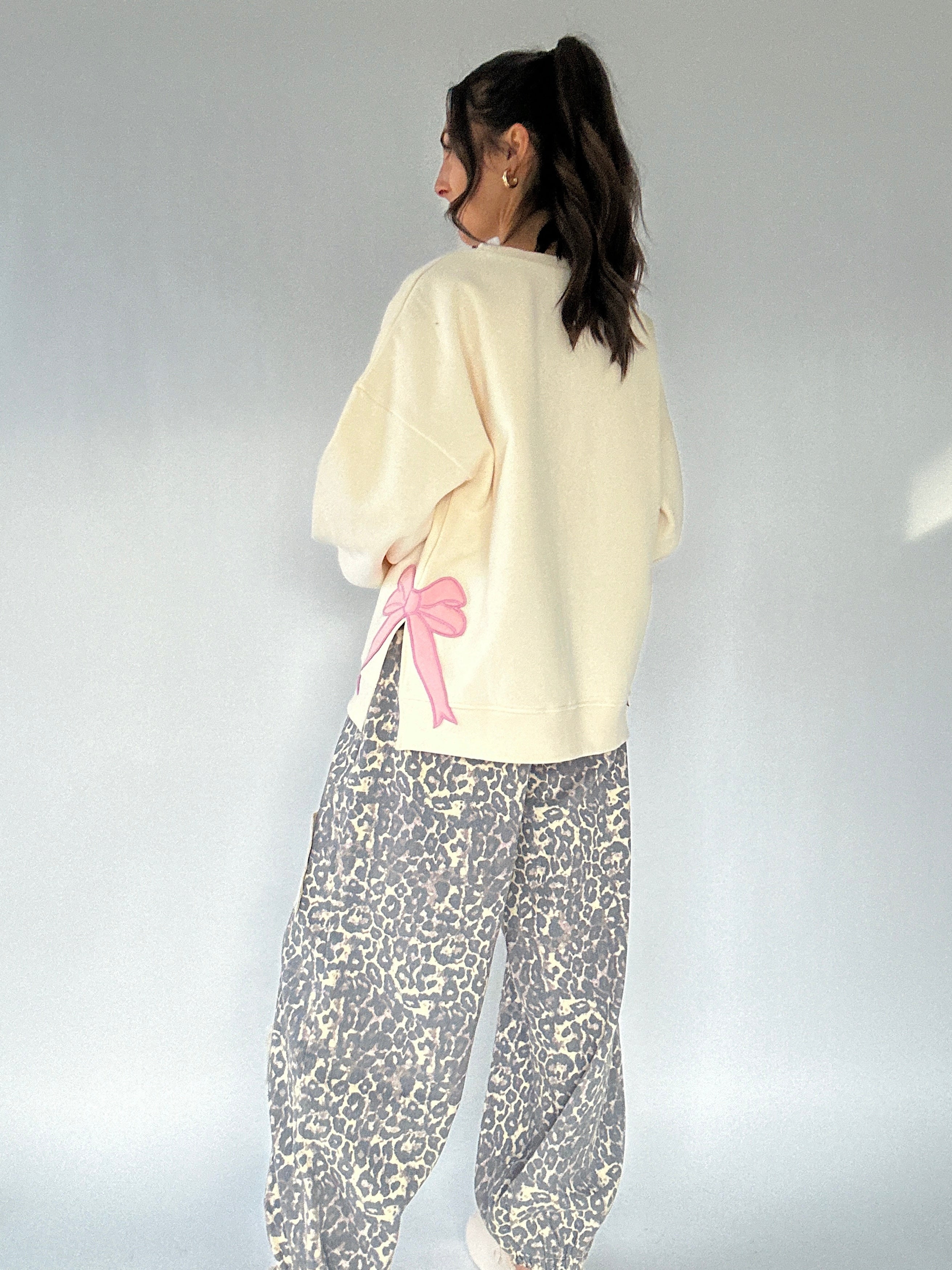 Person wearing a cream sweatshirt with a pink bow and leopard patterned pants on a plain background