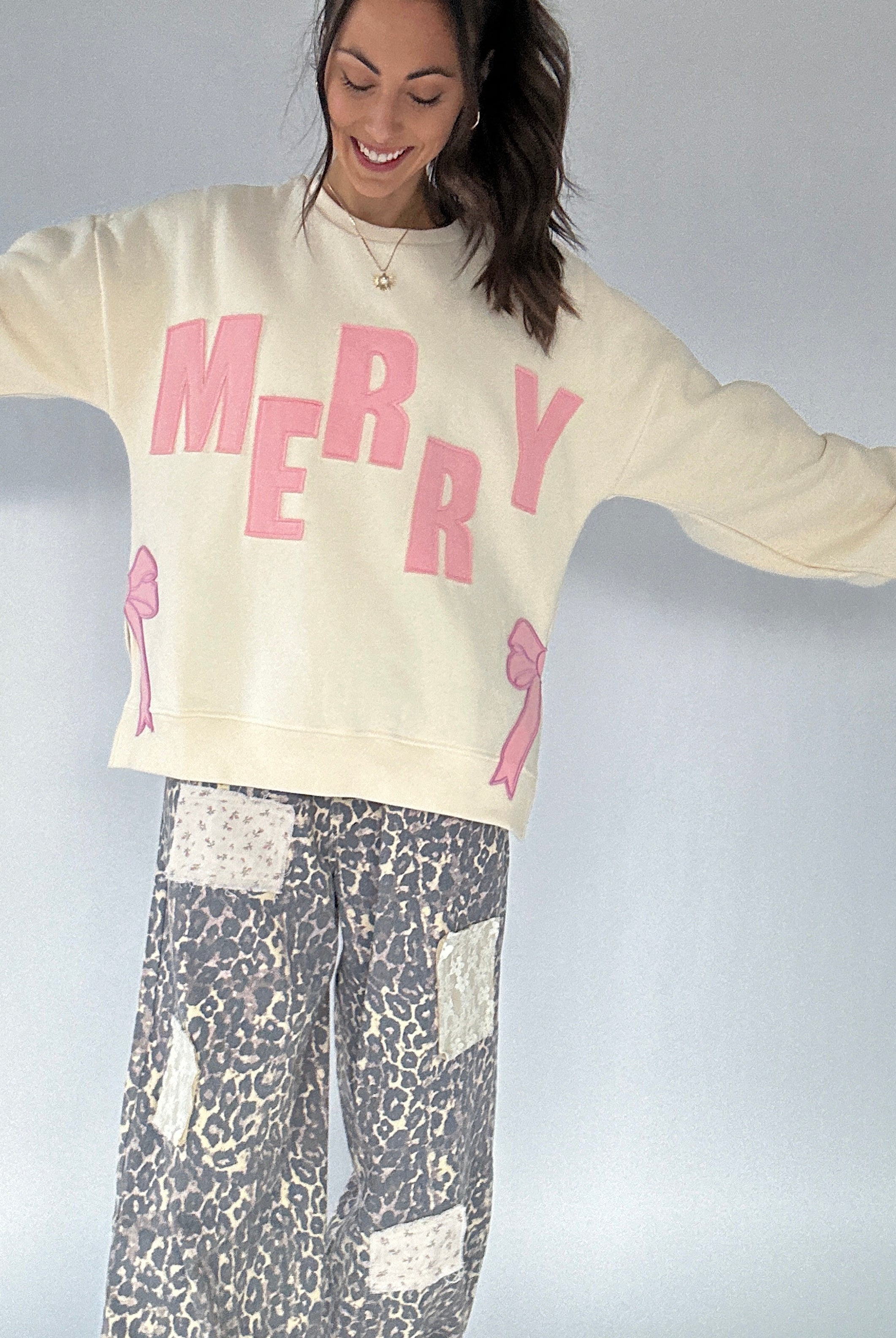 Person wearing a cream sweatshirt with 'MERRY' in pink and leopard patterned pants on a plain background