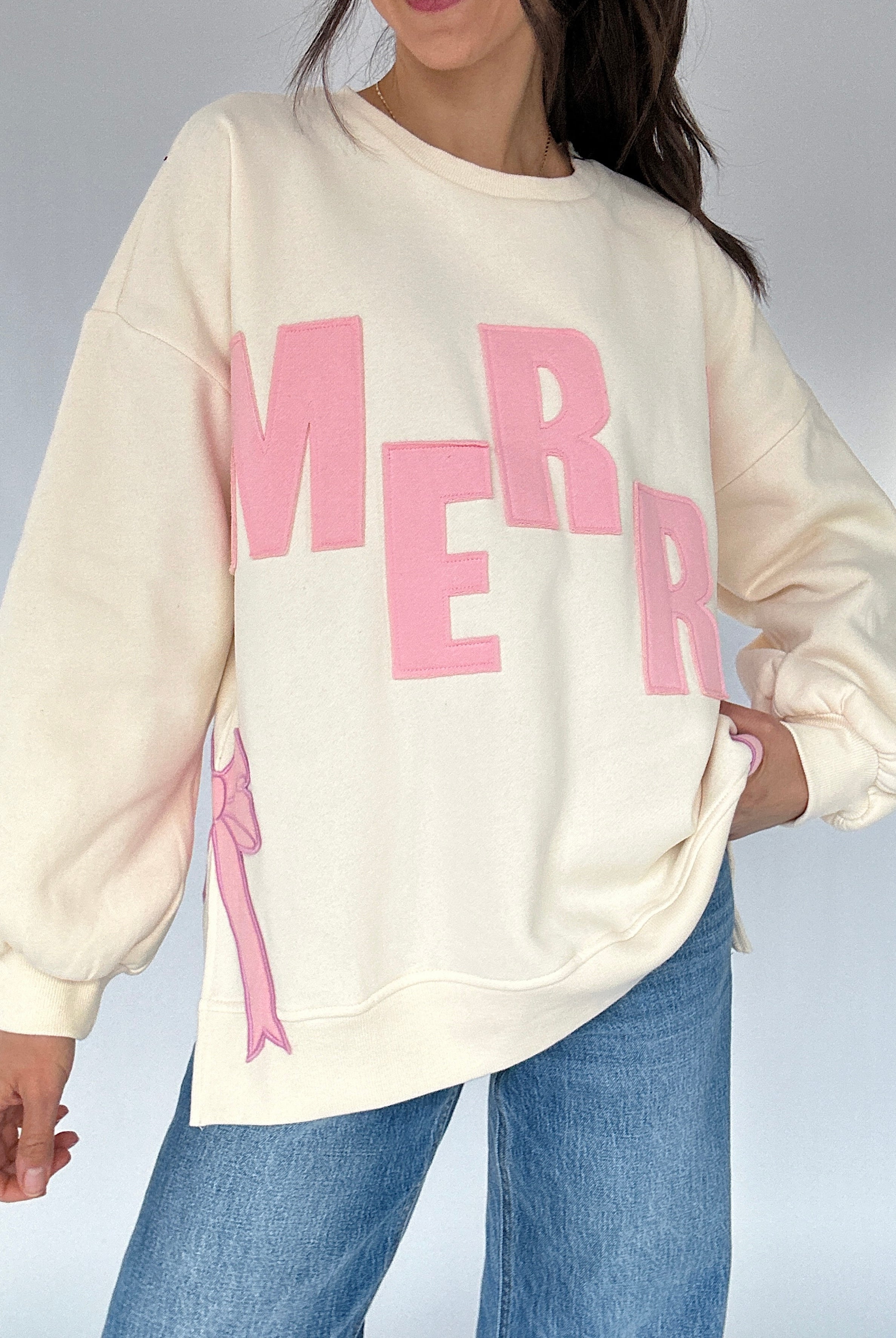 Person wearing a cream sweatshirt with pink text and a pink bow design, standing against a neutral background.