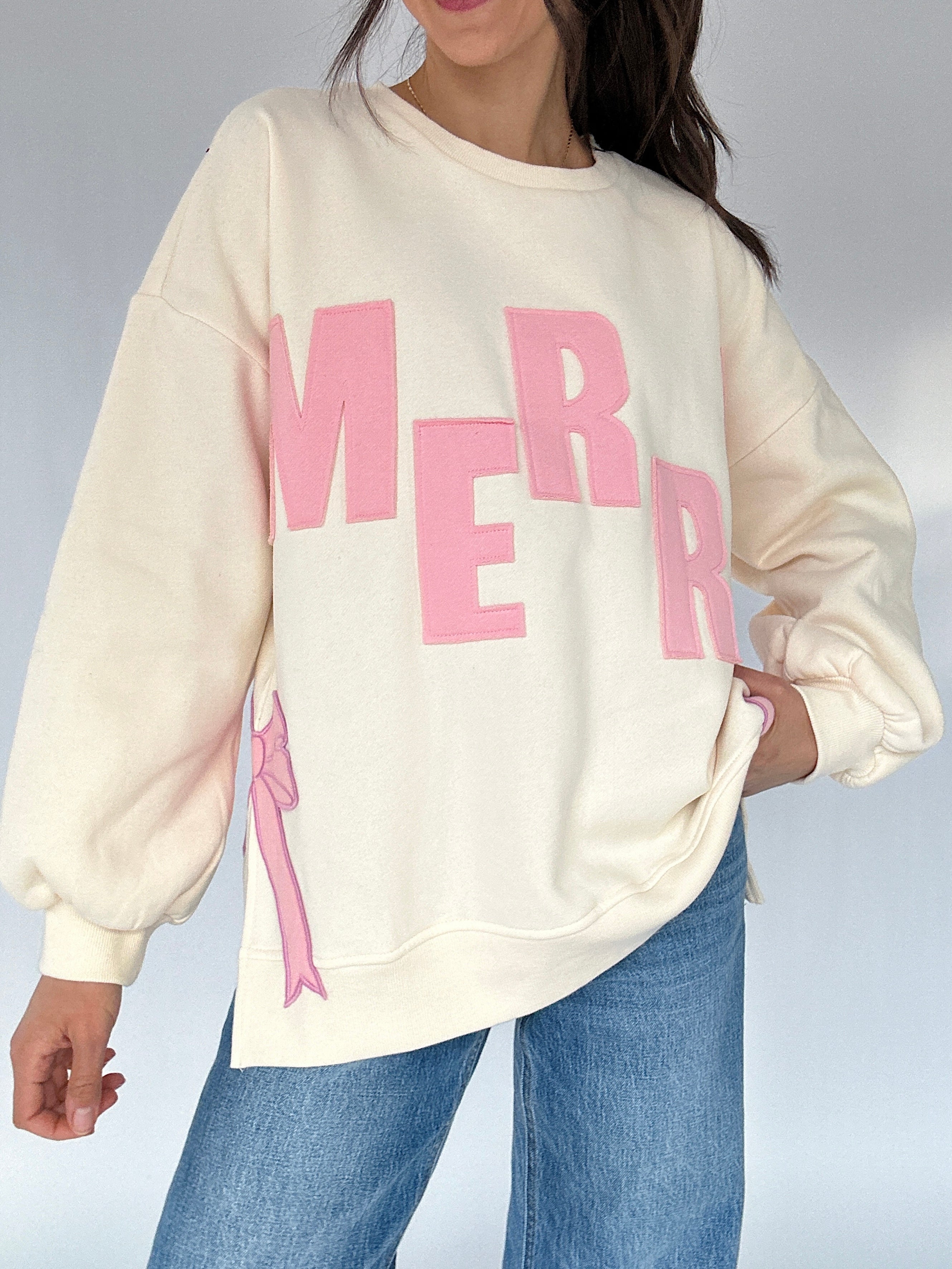 Person wearing a cream sweatshirt with pink text and a pink bow design, standing against a neutral background.