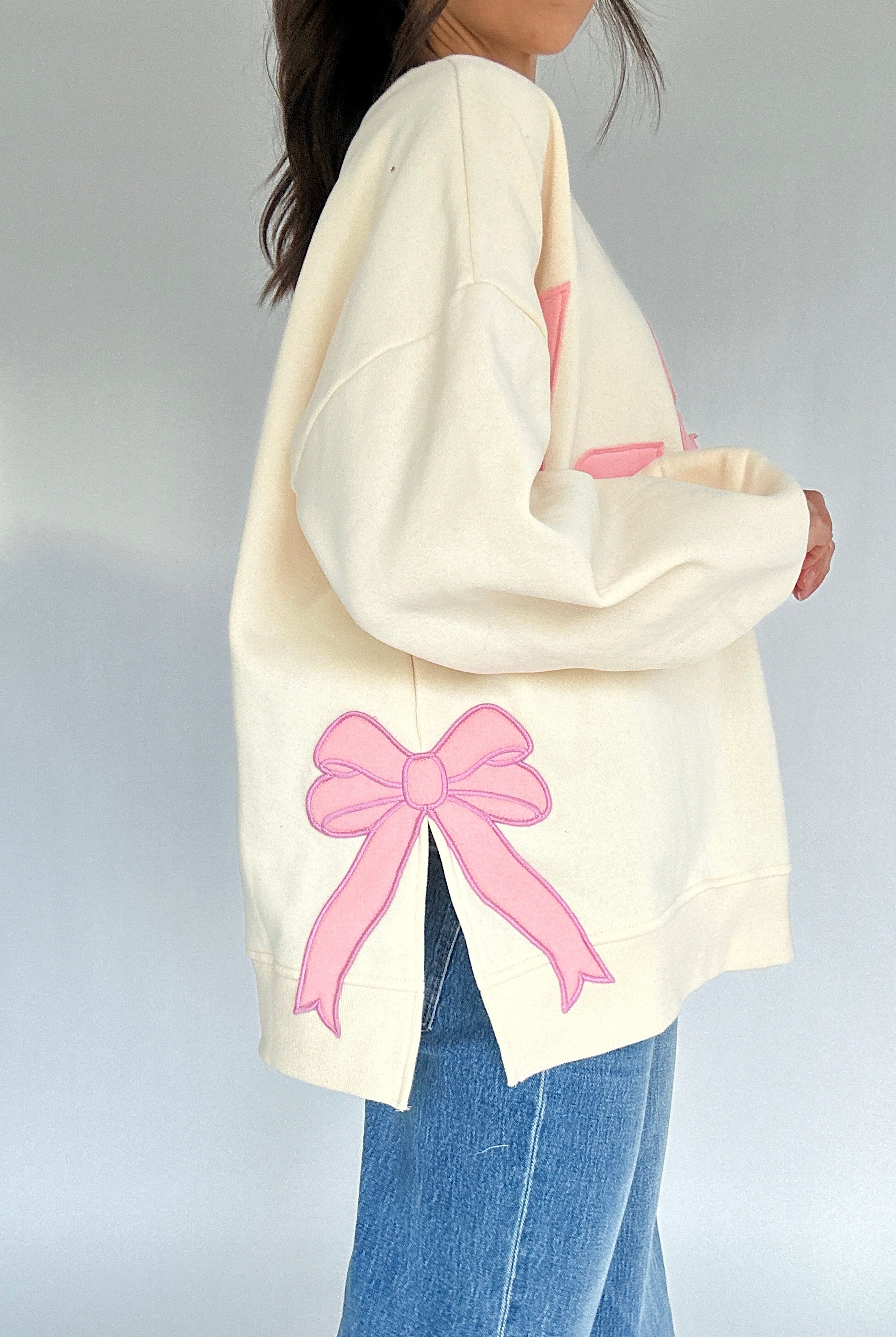 White sweatshirt with pink bow patch on side worn by a person on a plain background