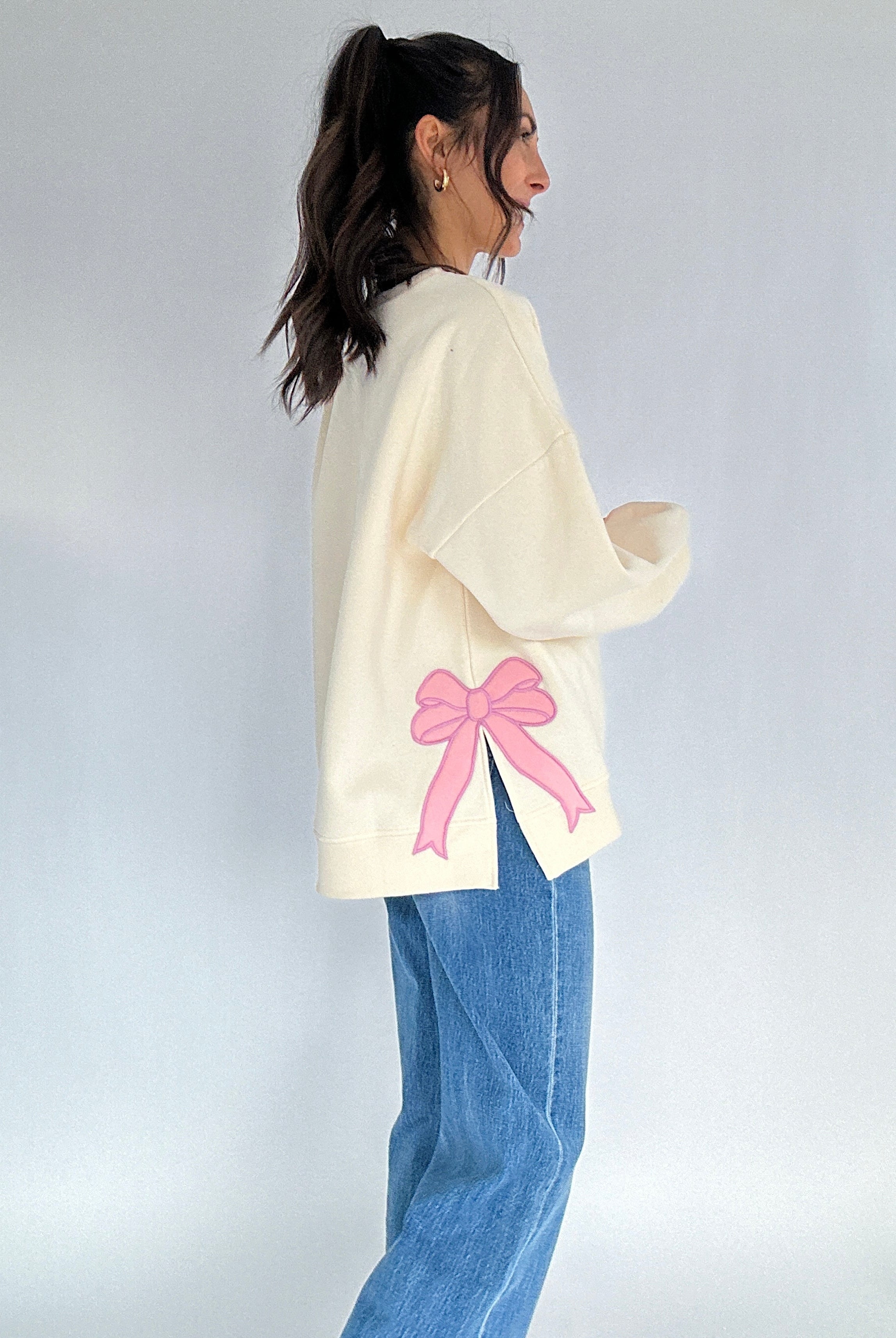 Person wearing a cream sweater with a pink bow patch on side on a plain background