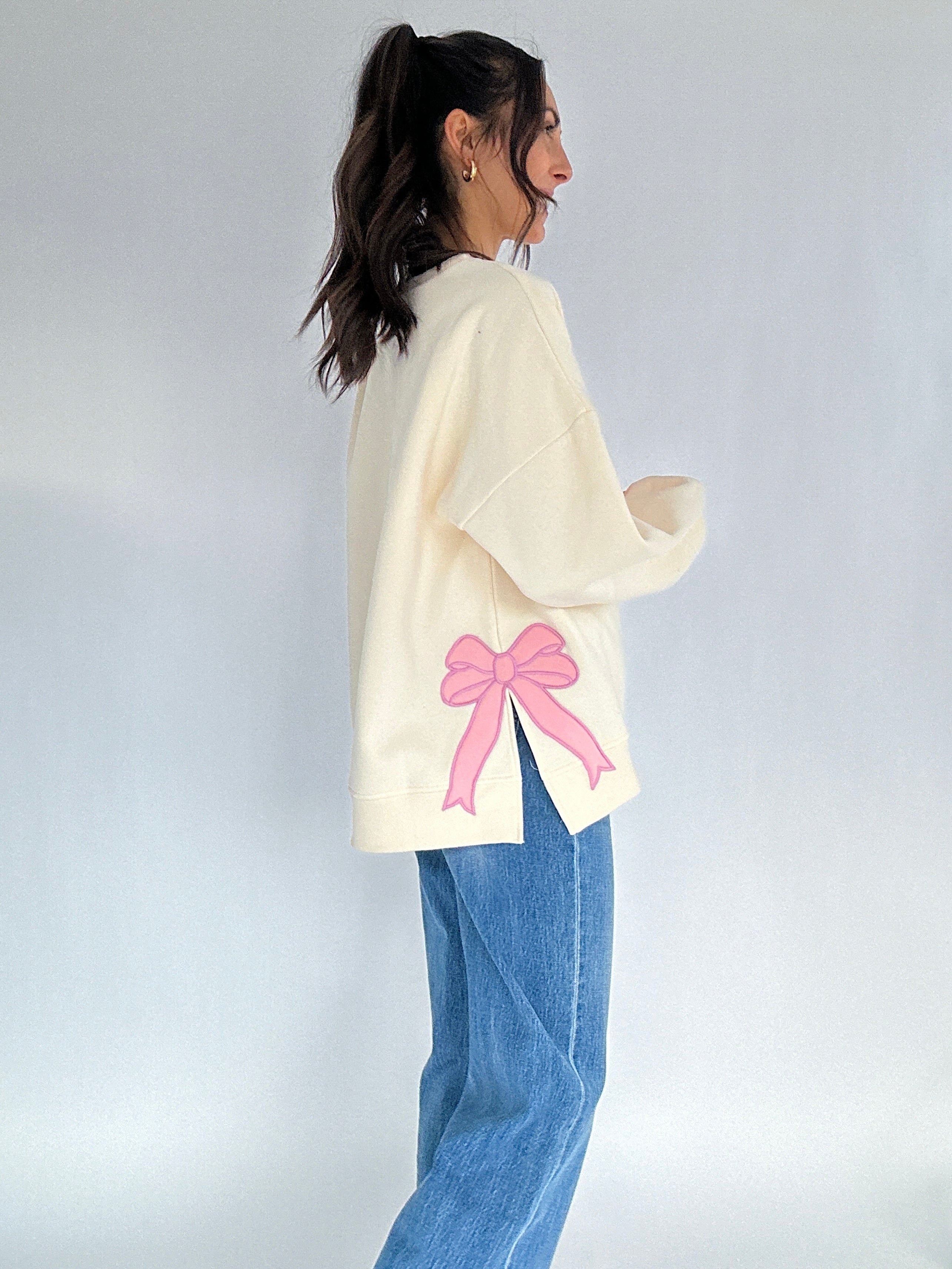 Person wearing a cream sweater with a pink bow patch on side on a plain background