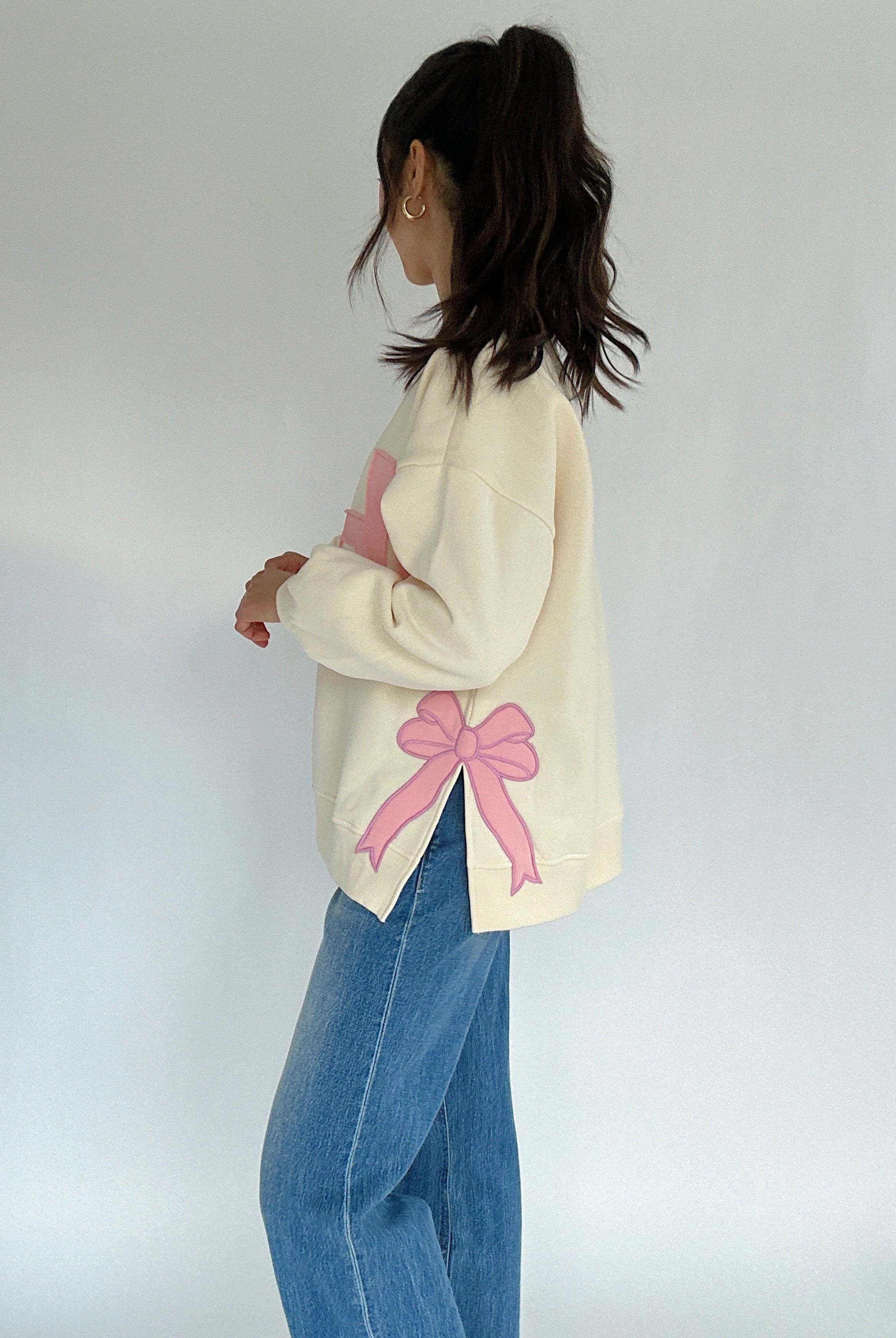 Person wearing a cream sweater with a pink bow patch on side and blue jeans on a white background