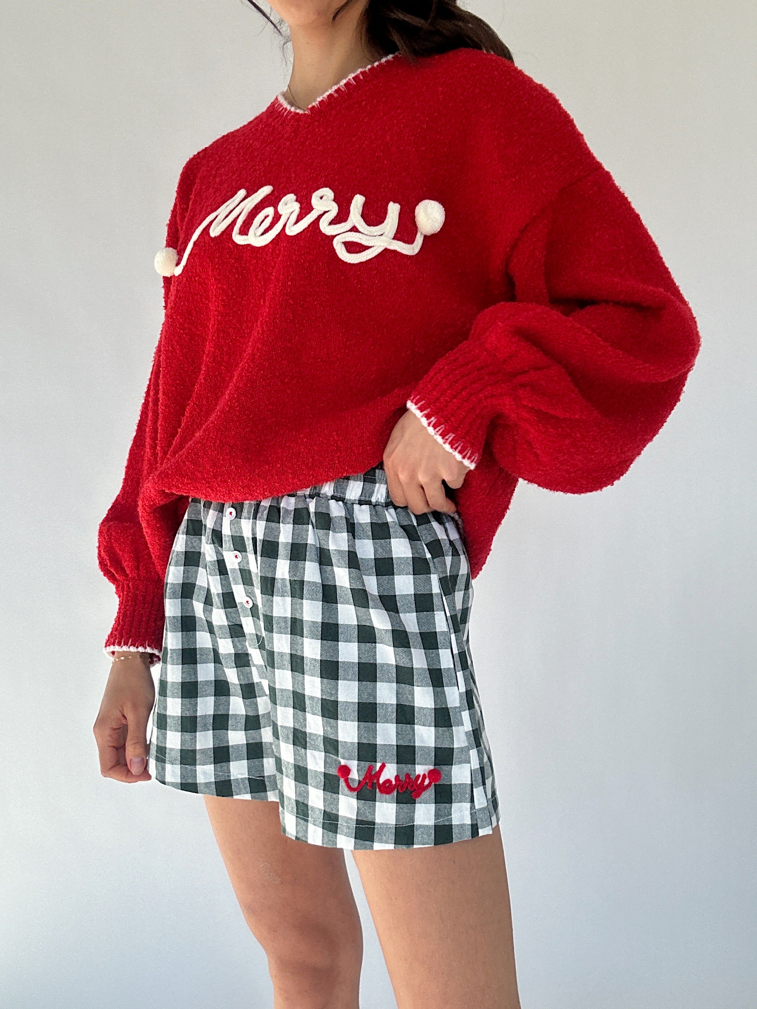Person wearing a red sweater with 'Merry' text and checkered shorts with matching text on a plain background to show details