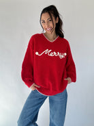 Person wearing a red v-neck sweater with white 'Merry' text with jeans on a plain background