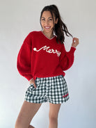 Person wearing a red sweater with white 'Merry' text and green checkered shorts on a plain background