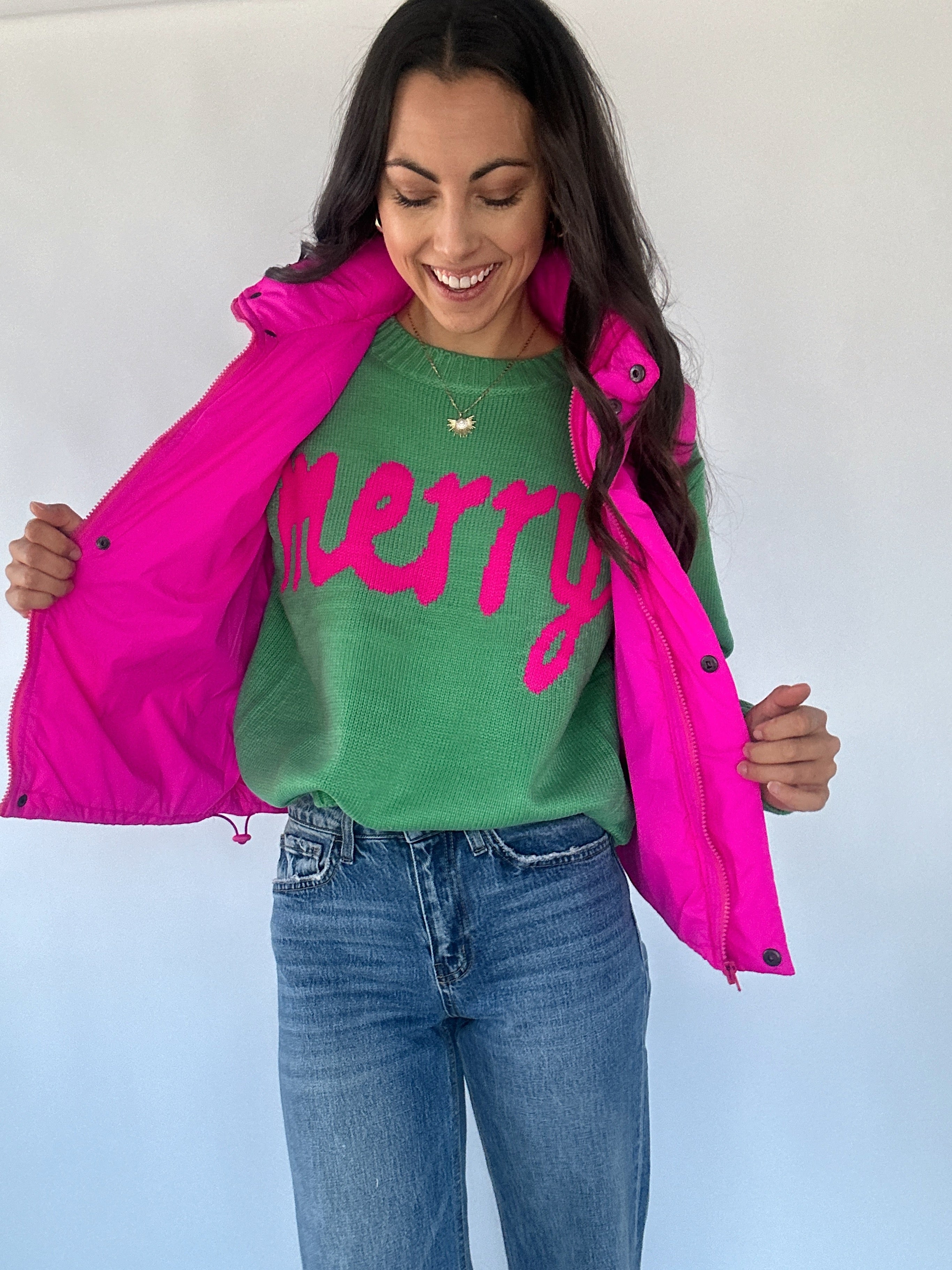 Woman wearing a green sweater with pink 'merry' lettering and a pink puffer vest on a plain background