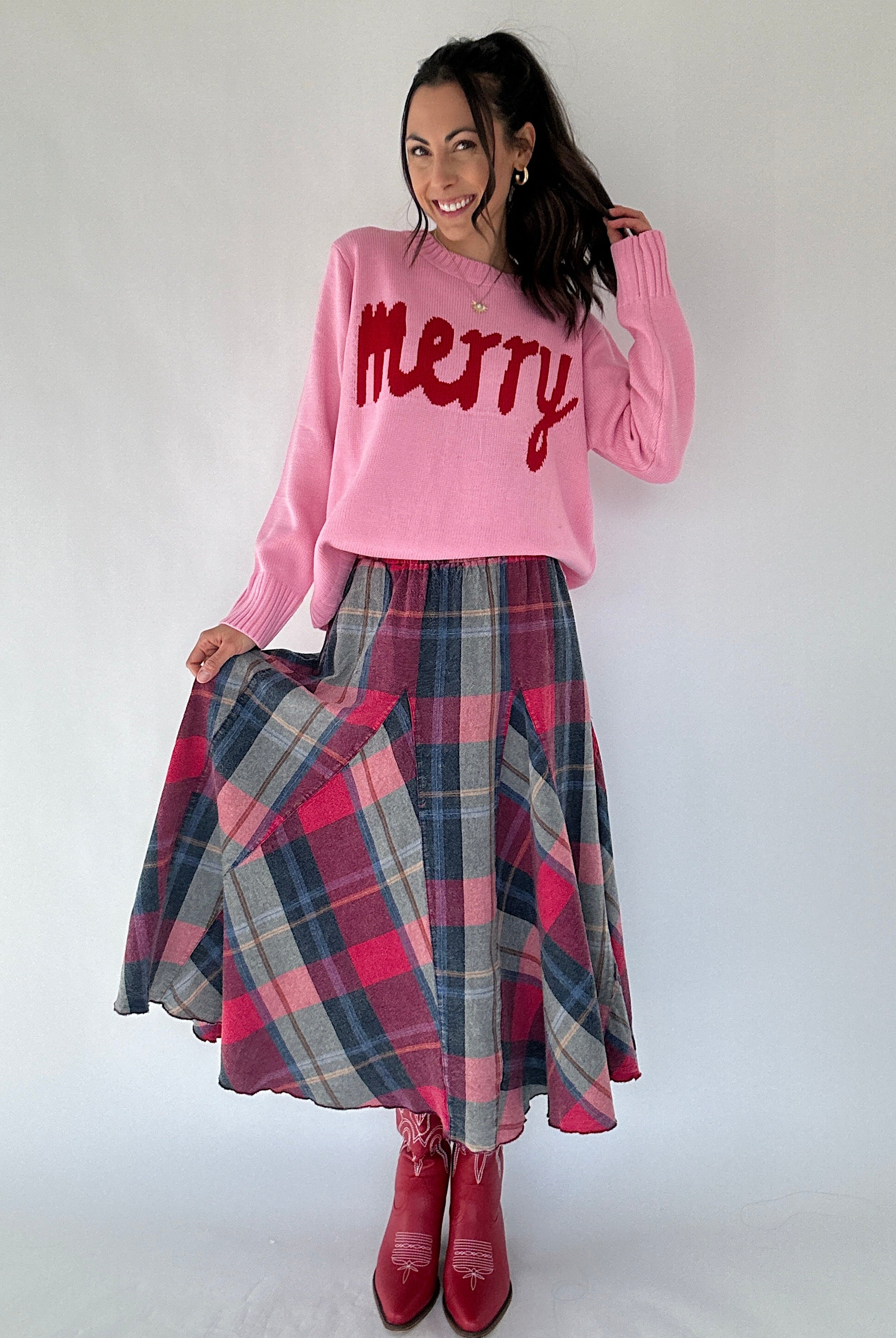 Woman wearing a pink sweater with 'Merry' text and a plaid skirt on a white background