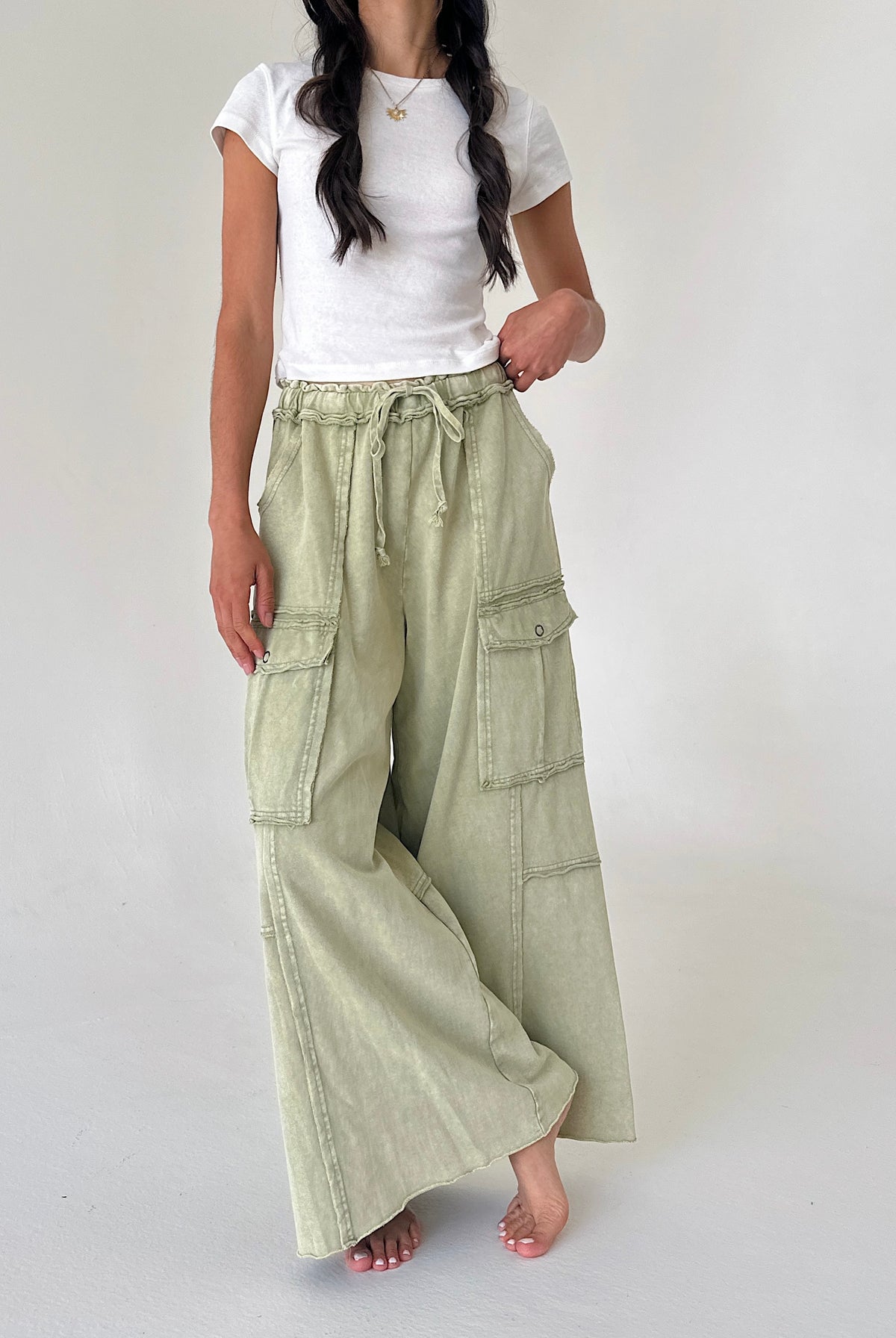 A woman wearing wide-leg sage green cargo pants with a drawstring waist, styled with a white t-shirt, standing against a plain light gray studio background.