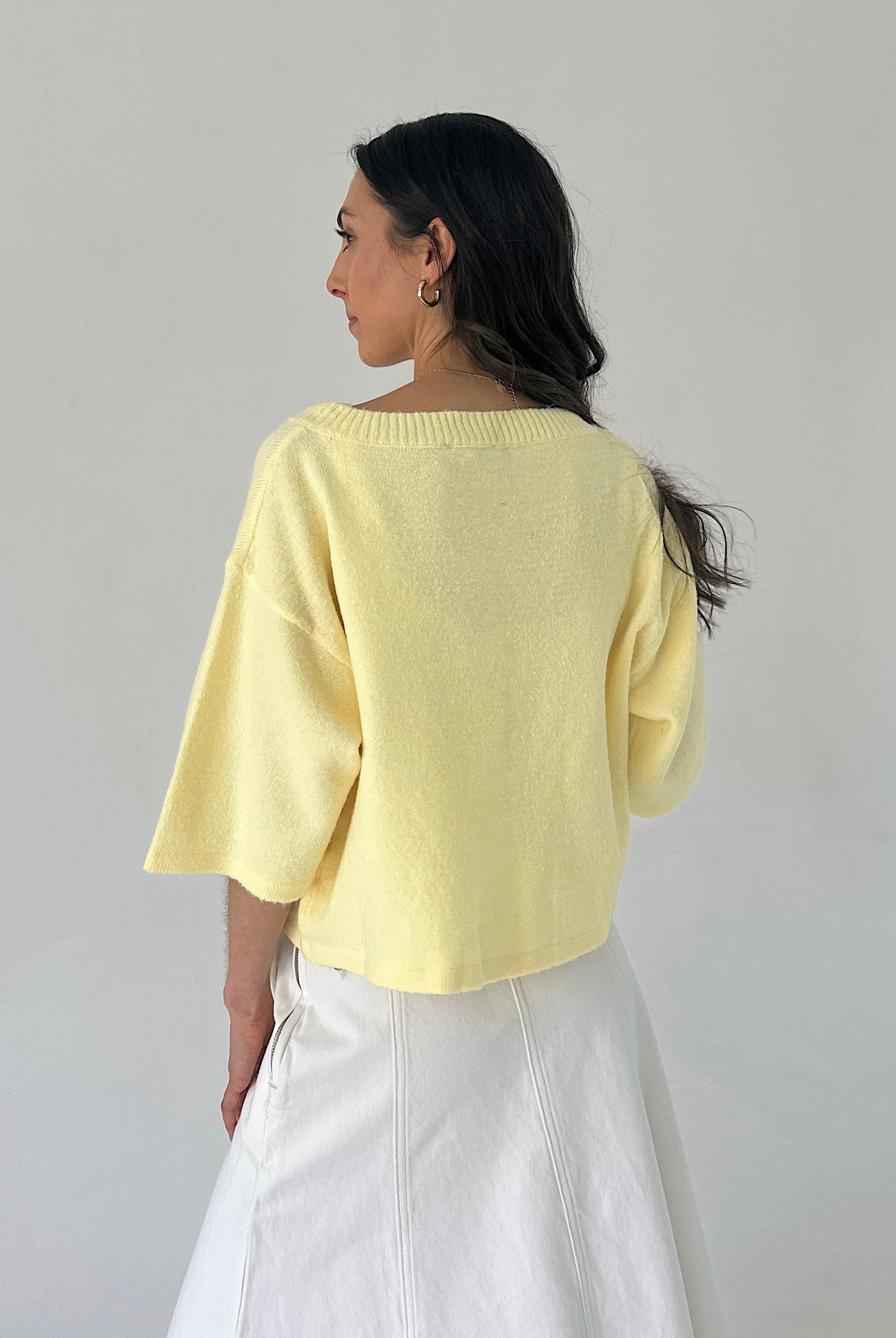 Woman wearing a light yellow sweater and white pants against a plain background showing back