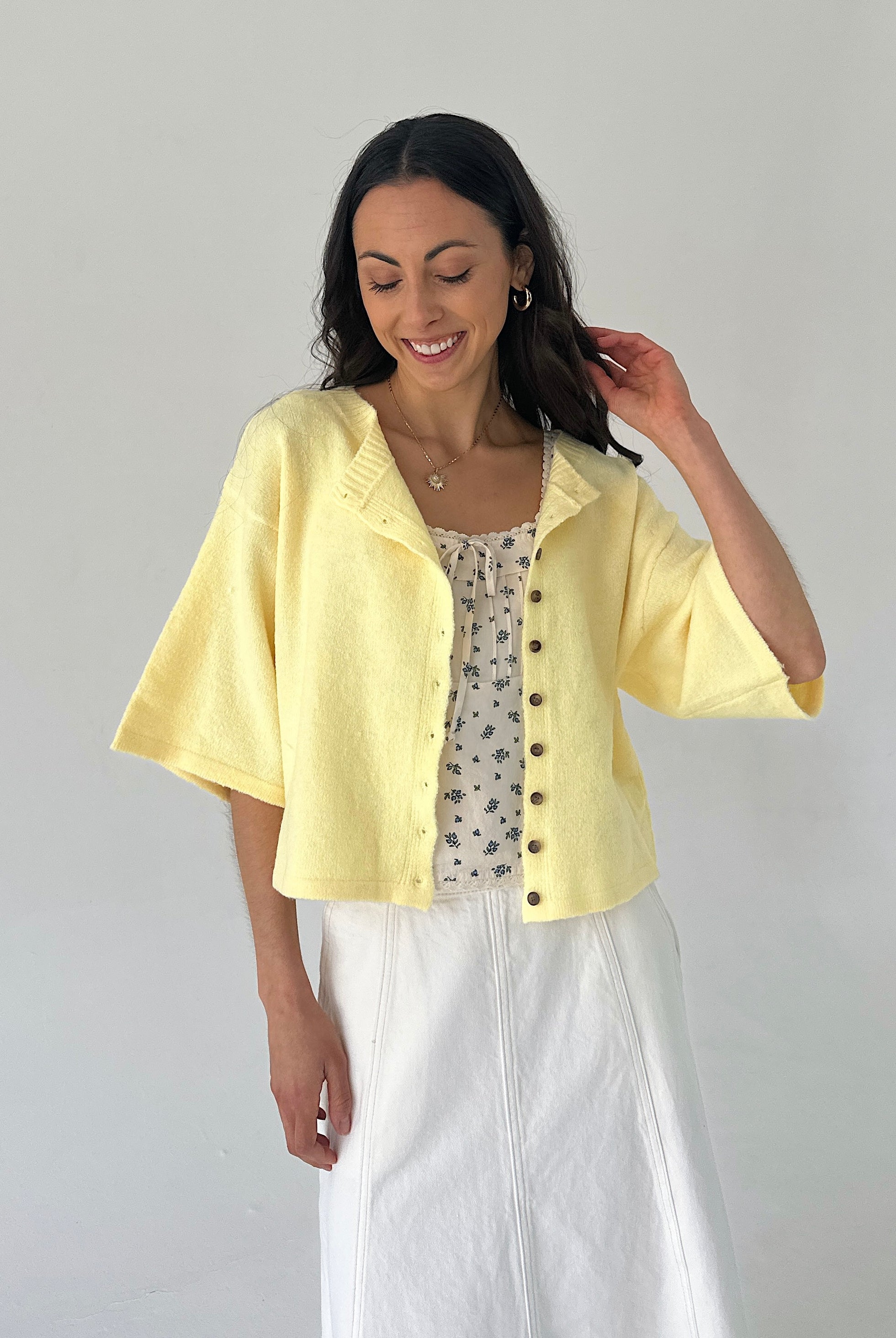 Woman wearing a light yellow cardigan over a patterned top and white skirt on a plain background