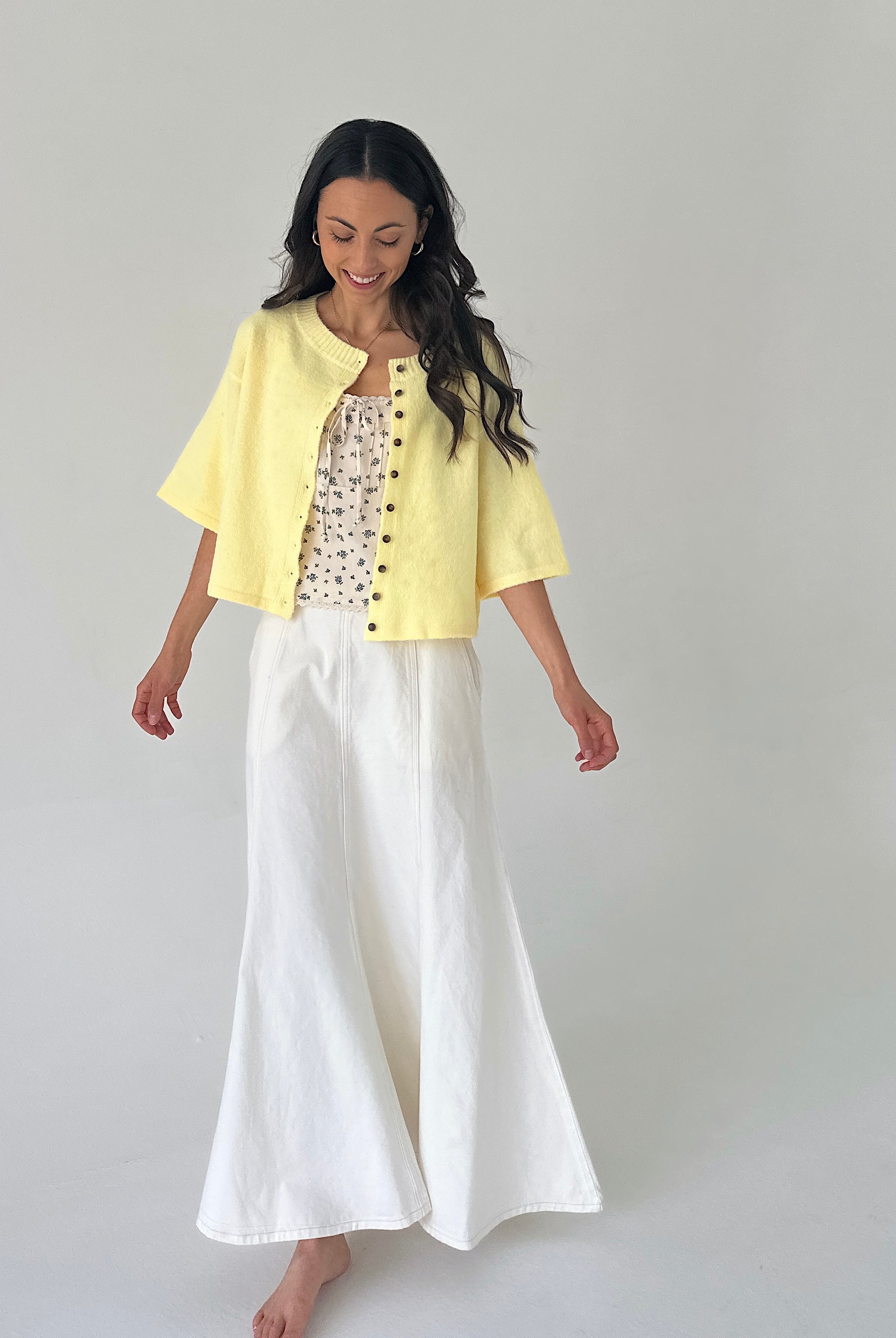 Woman wearing a yellow jacket over a patterned top and white skirt on a plain background