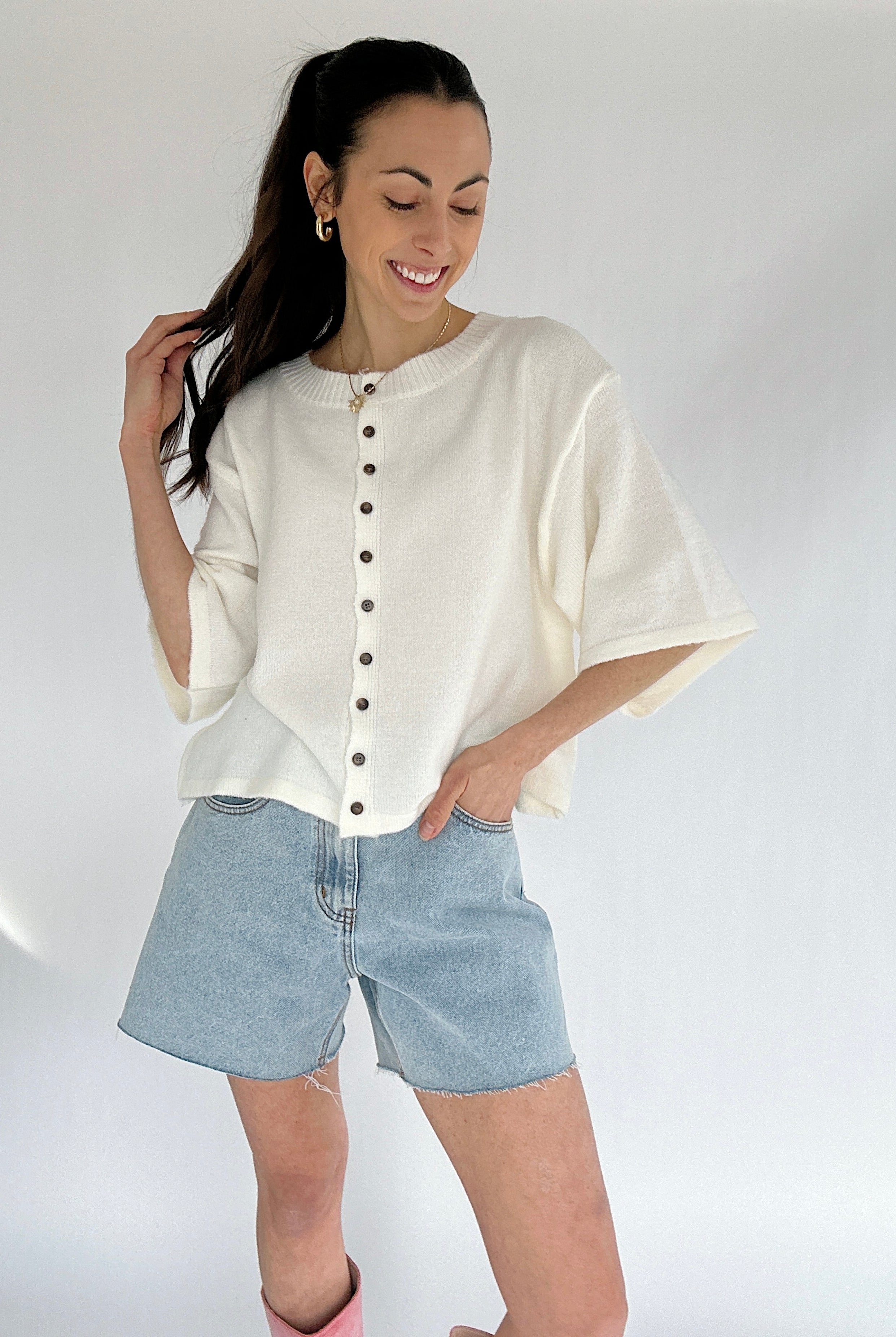 Woman wearing a white button-up short sleeve cardigan and denim shorts on a plain background