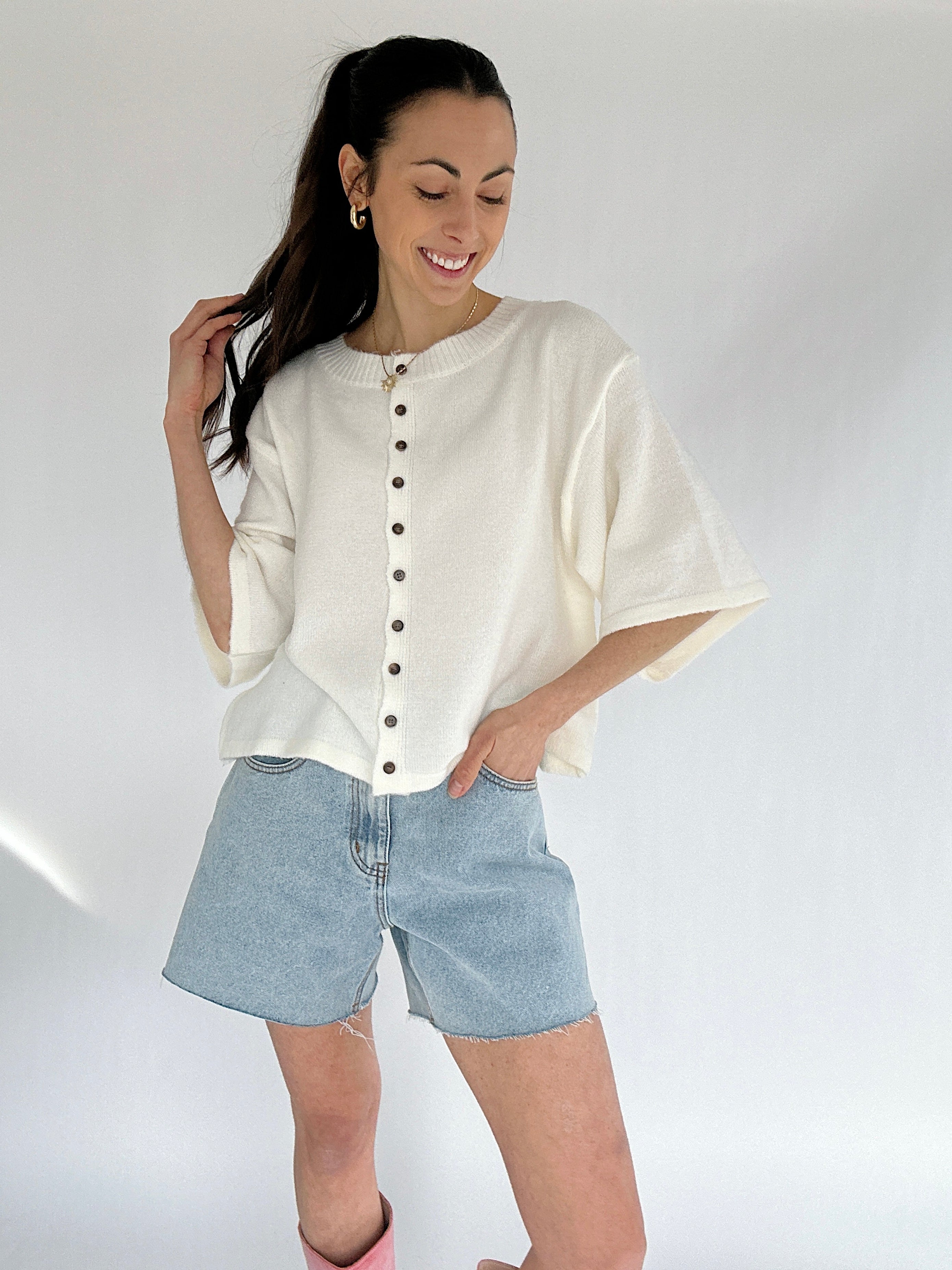 Woman wearing a white button-up short sleeve cardigan and denim shorts on a plain background