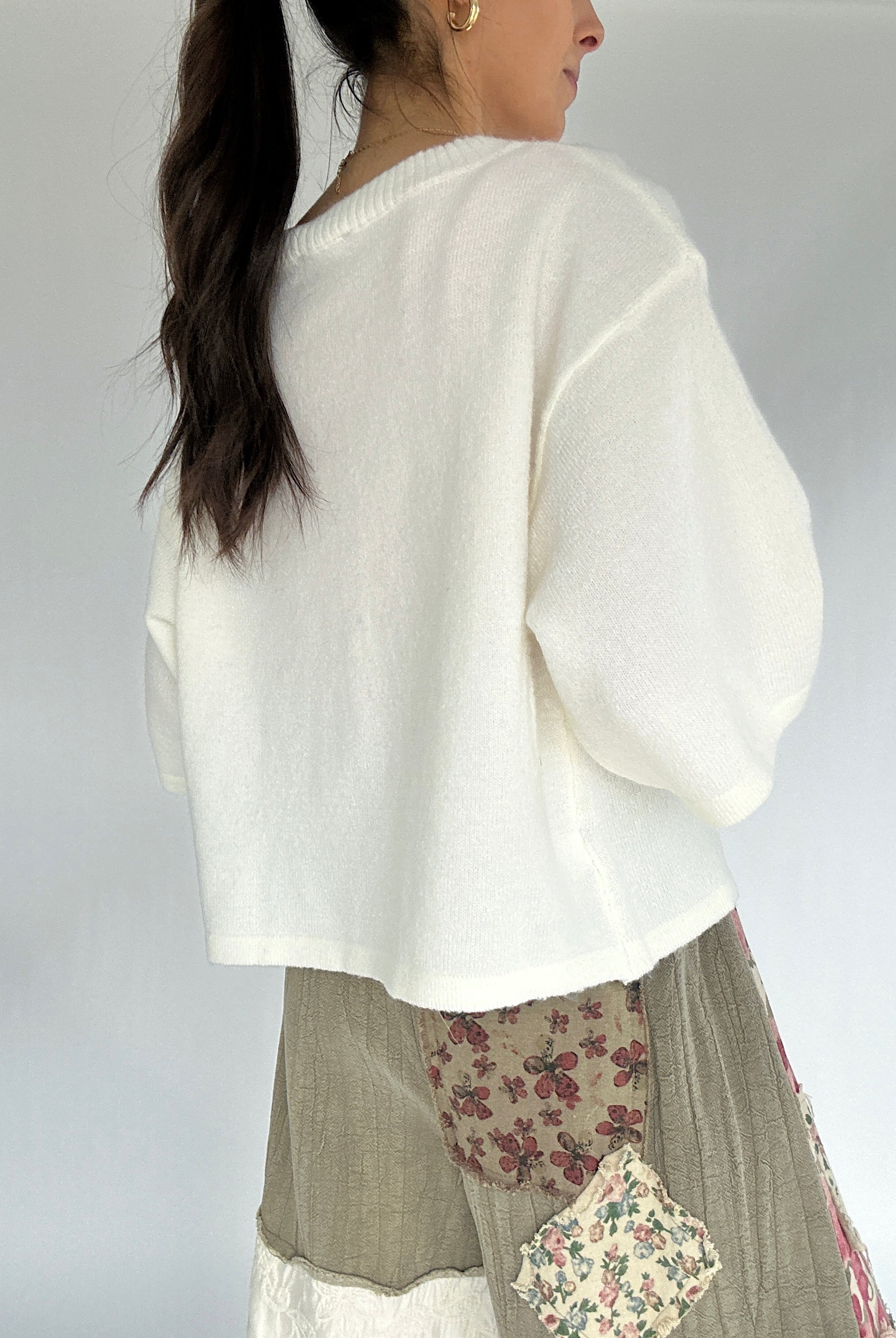Woman wearing a white sweater over a patterned skirt on a plain background from back