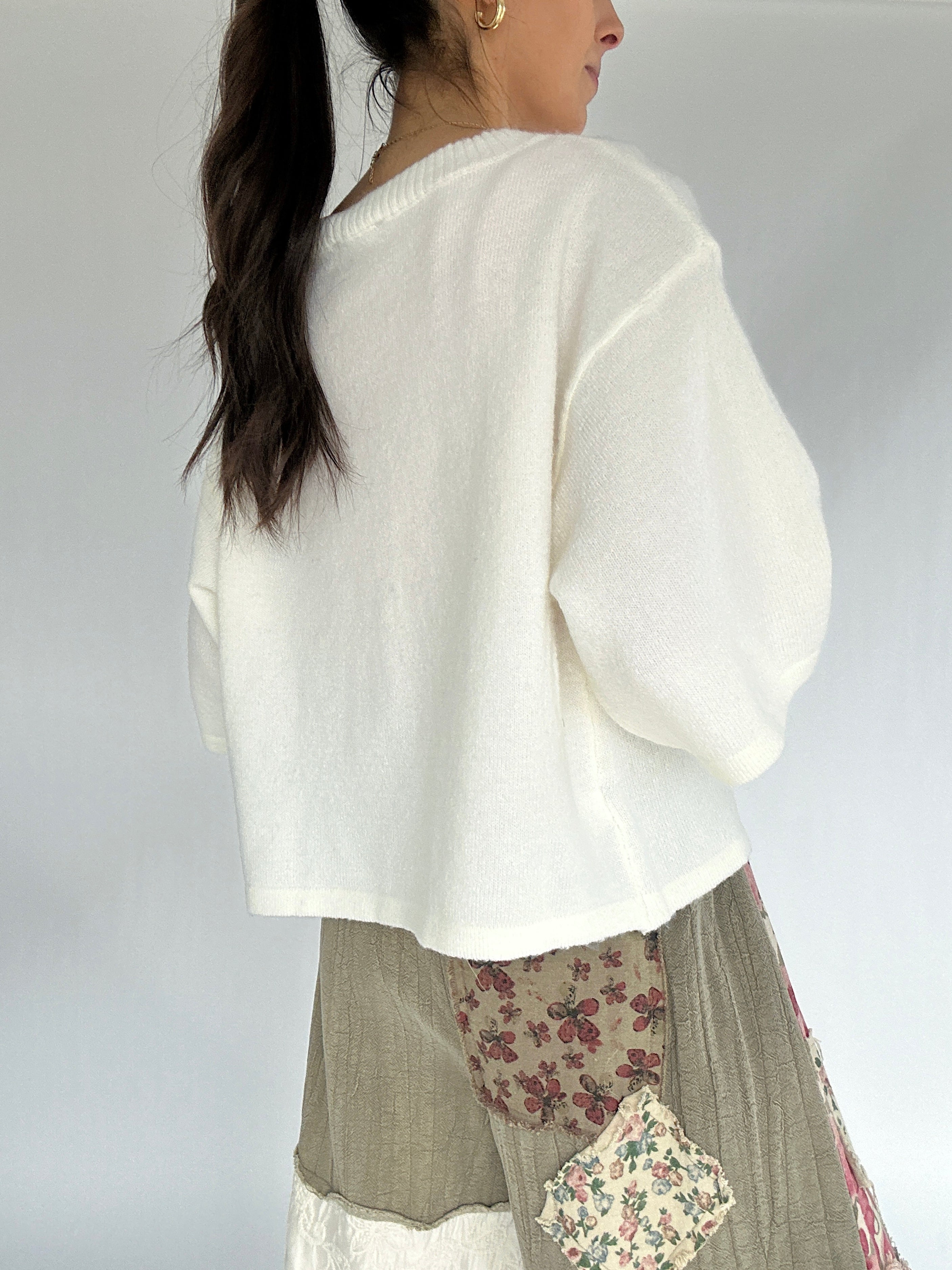 Woman wearing a white sweater over a patterned skirt on a plain background from back