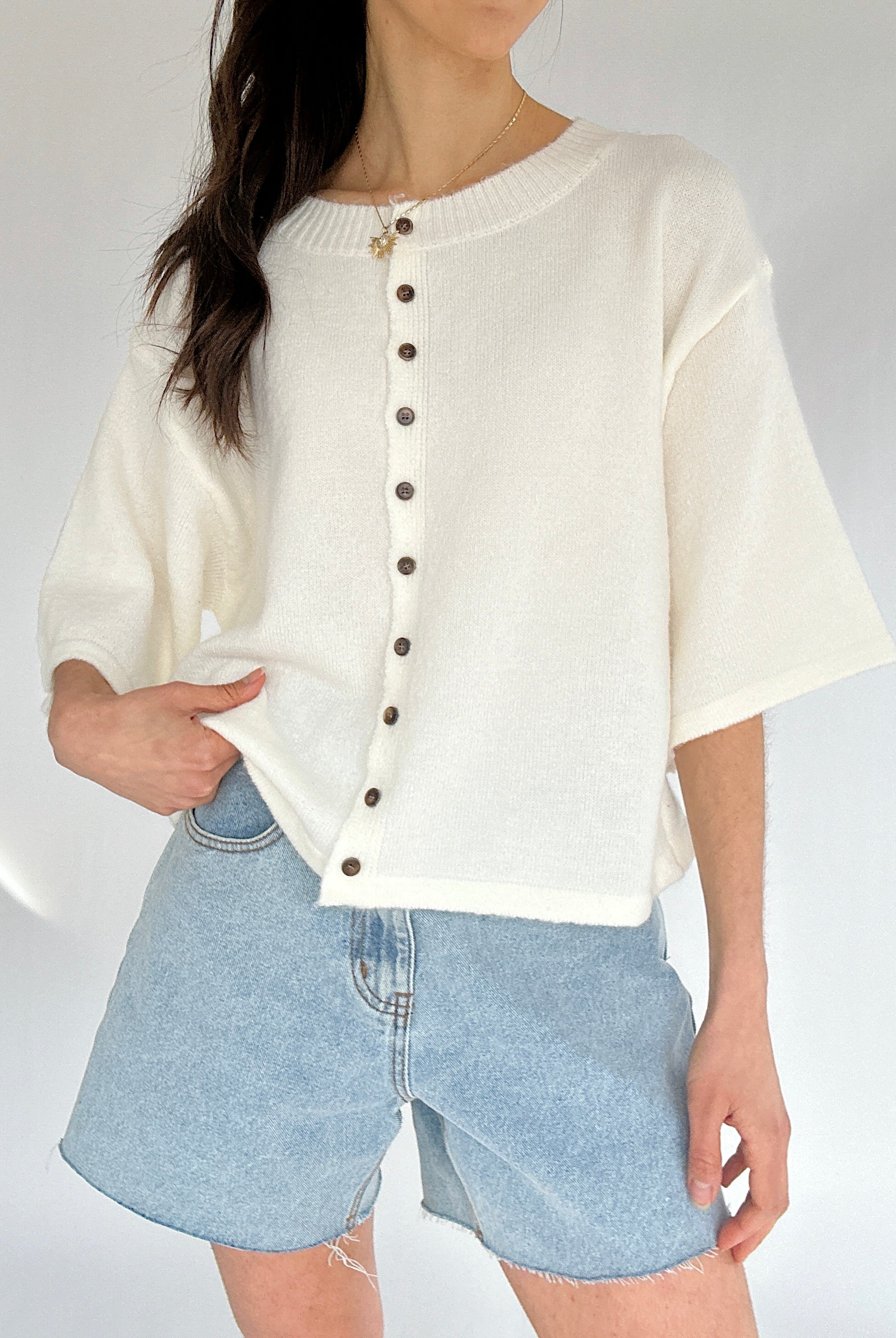 Person wearing a white button-up short sleeve cardigan and denim shorts on a plain background