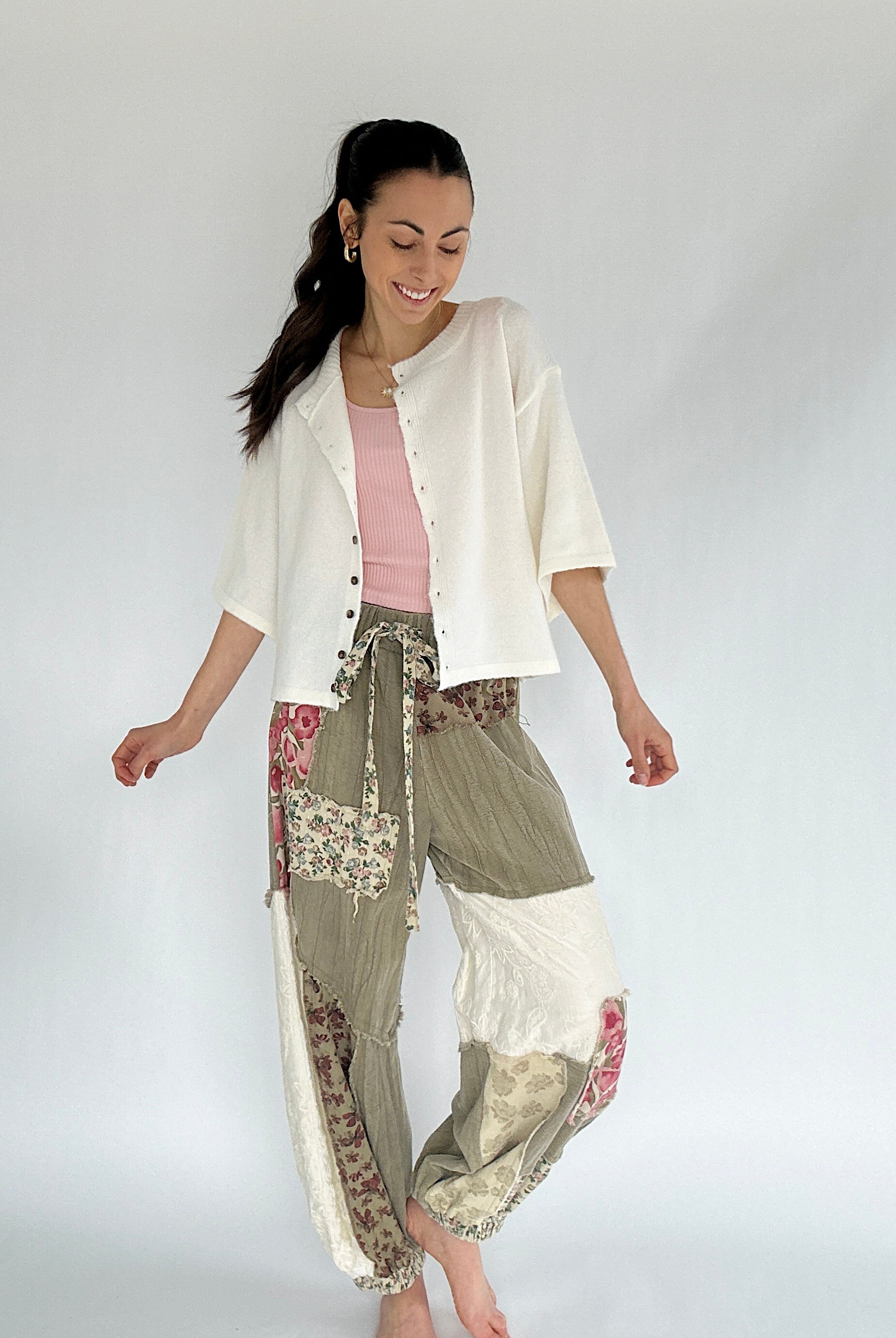 Woman wearing a white cardigan and olive floral patterned patchwork pants on a plain background