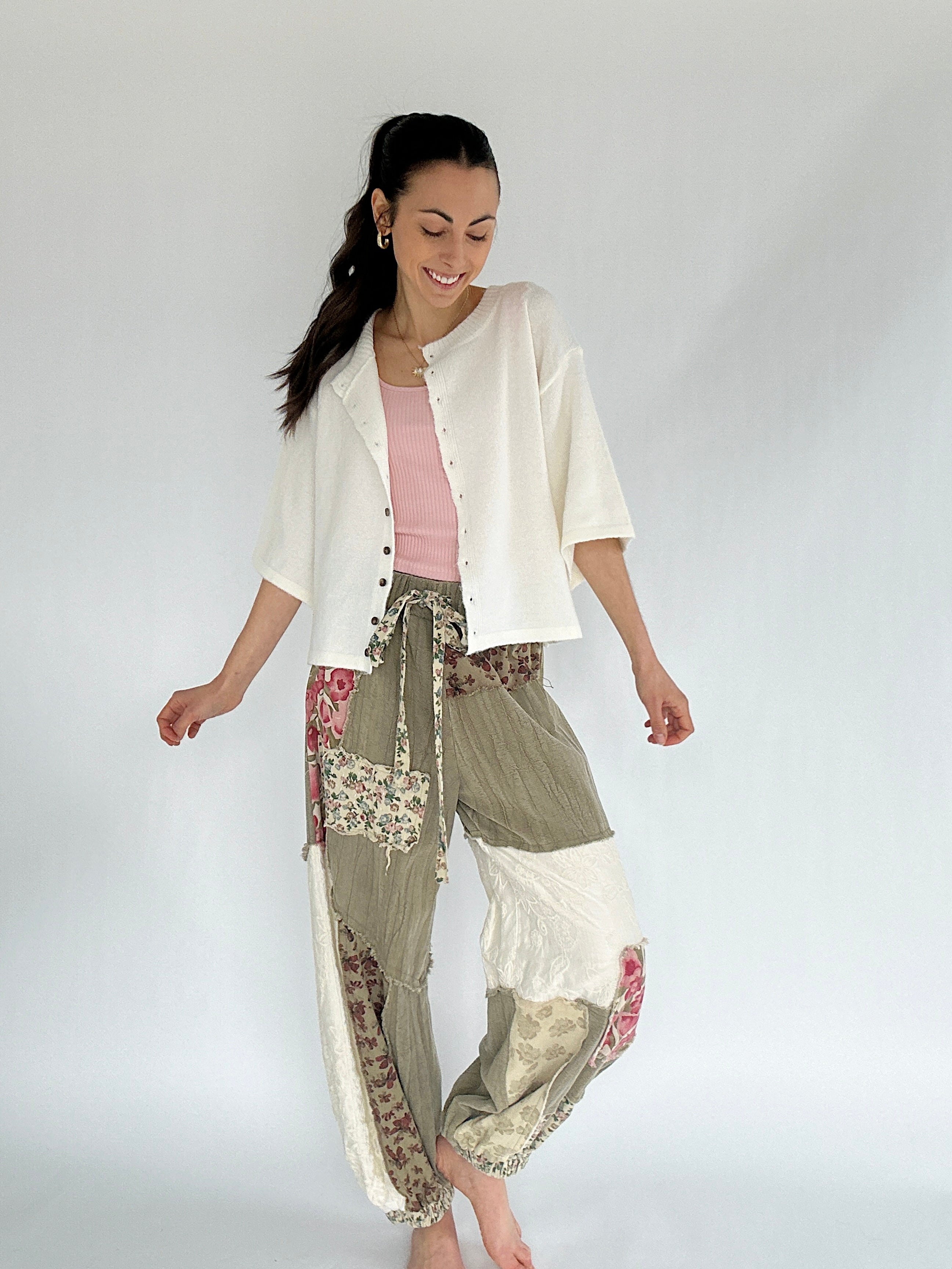 Woman wearing a white cardigan and olive floral patterned patchwork pants on a plain background