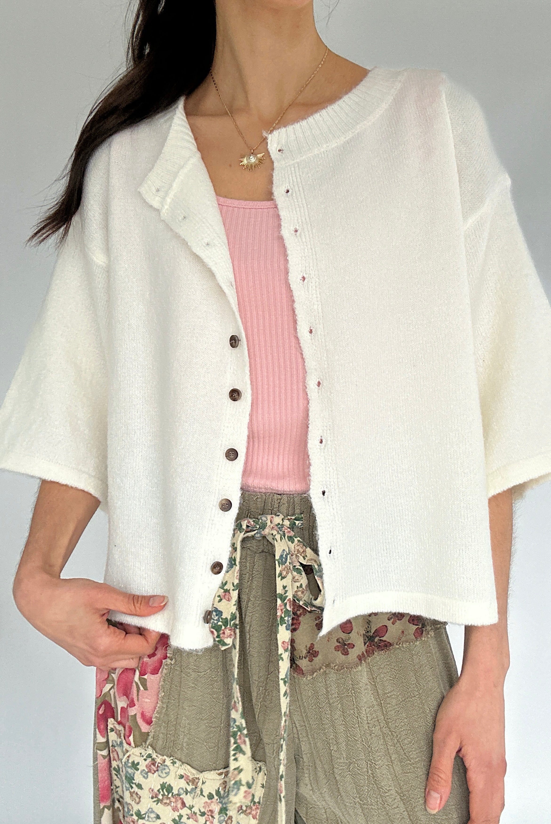 Person wearing a white cardigan over a pink top and floral pants on a gray background