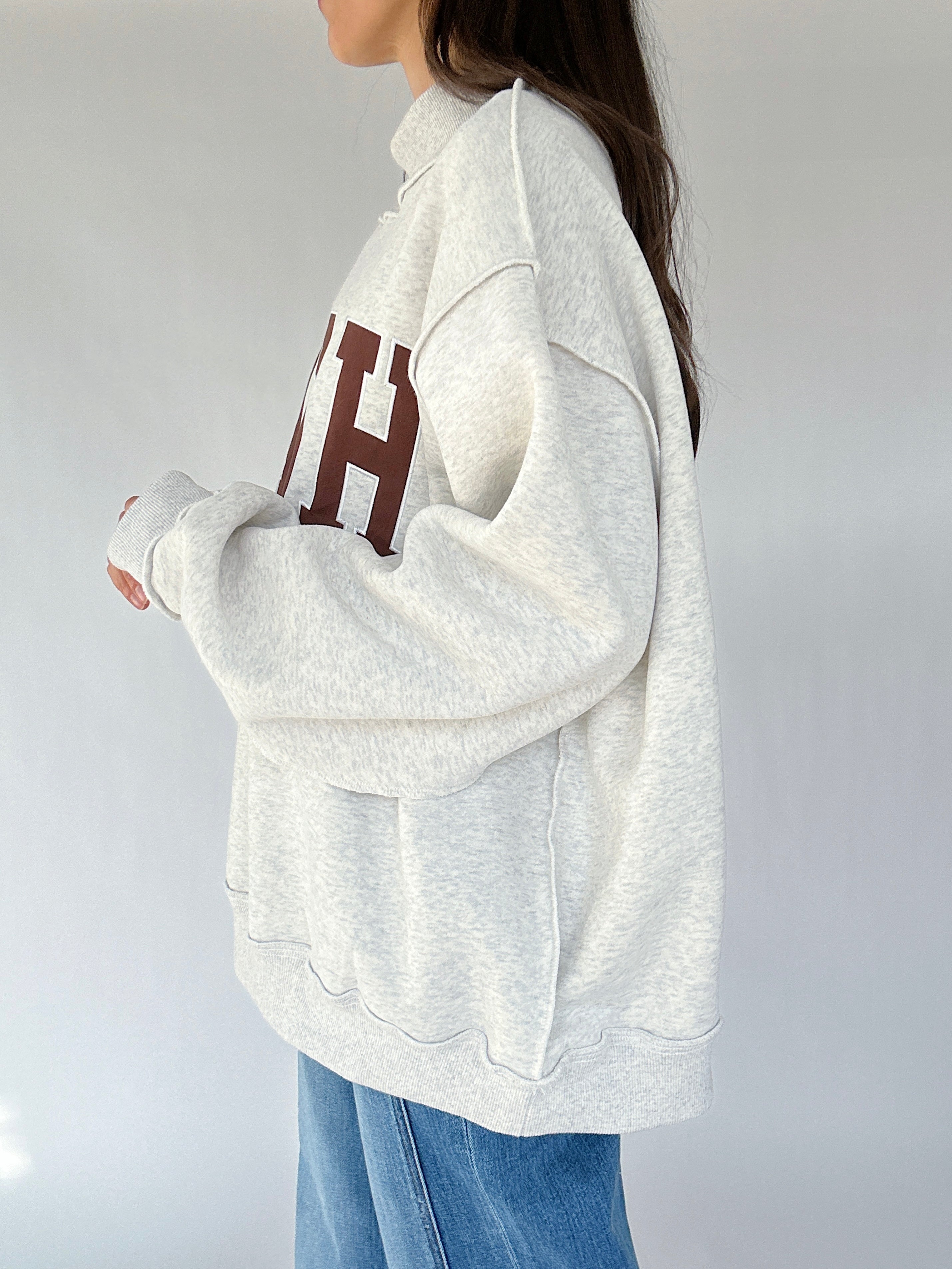 Person wearing a light gray sweatshirt with a brown logo on a plain background