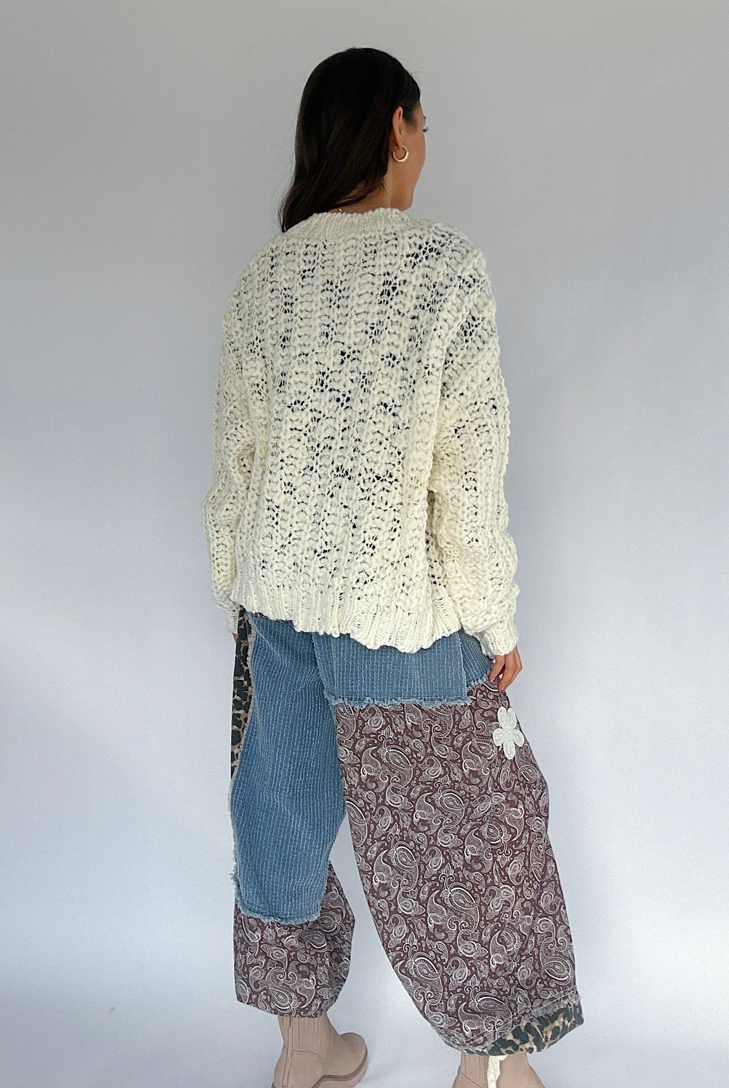 Person wearing a cream knitted cardigan, blue jeans, and patterned pants against a white background from back
