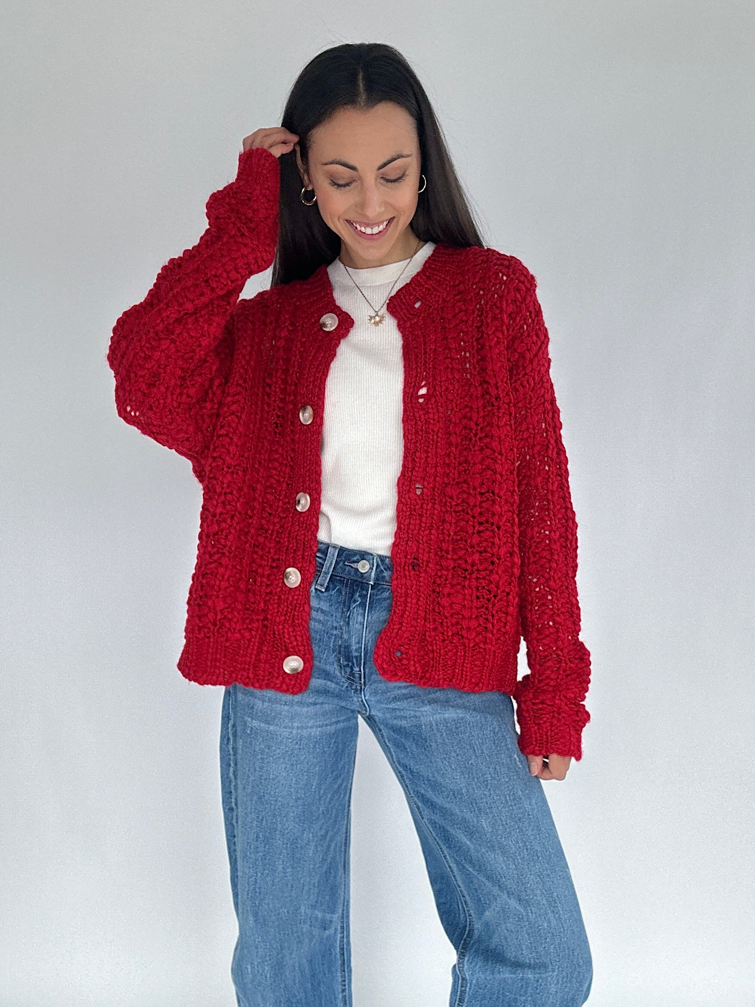 Woman wearing a red knitted cardigan over a white shirt and blue jeans on a plain background