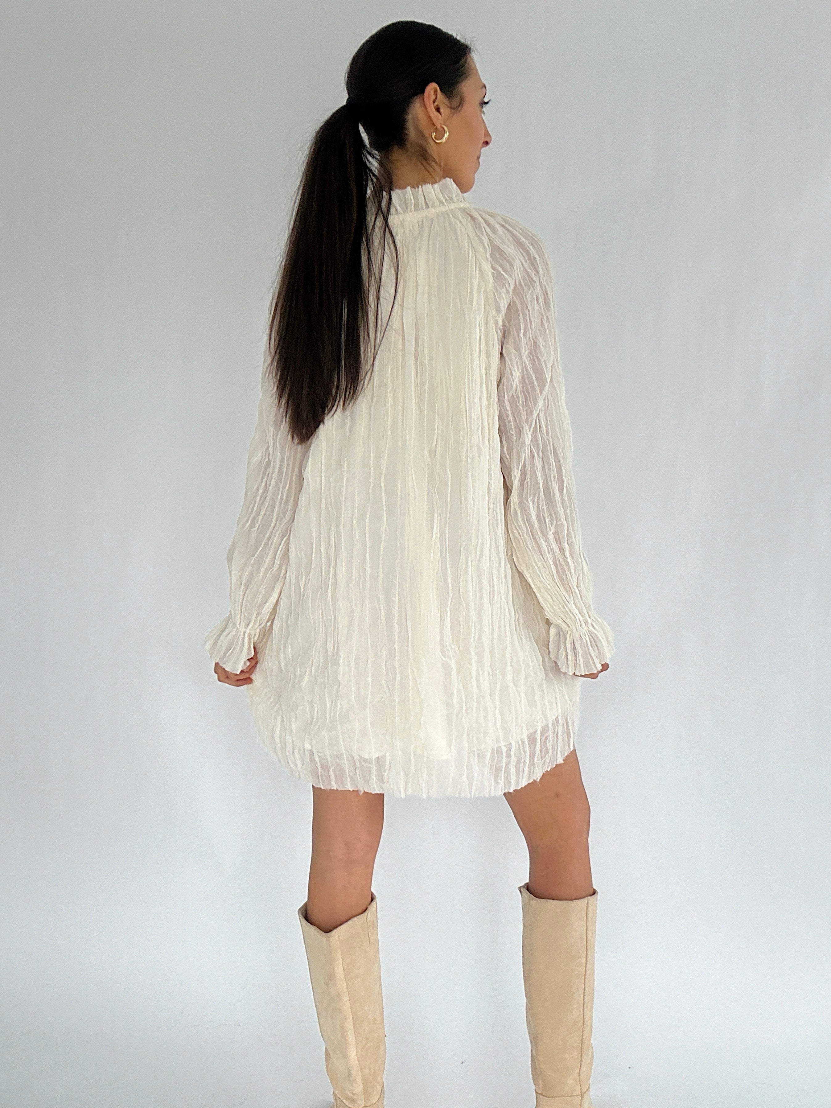Woman wearing a white pleated dress with long sleeves on a plain background showing back