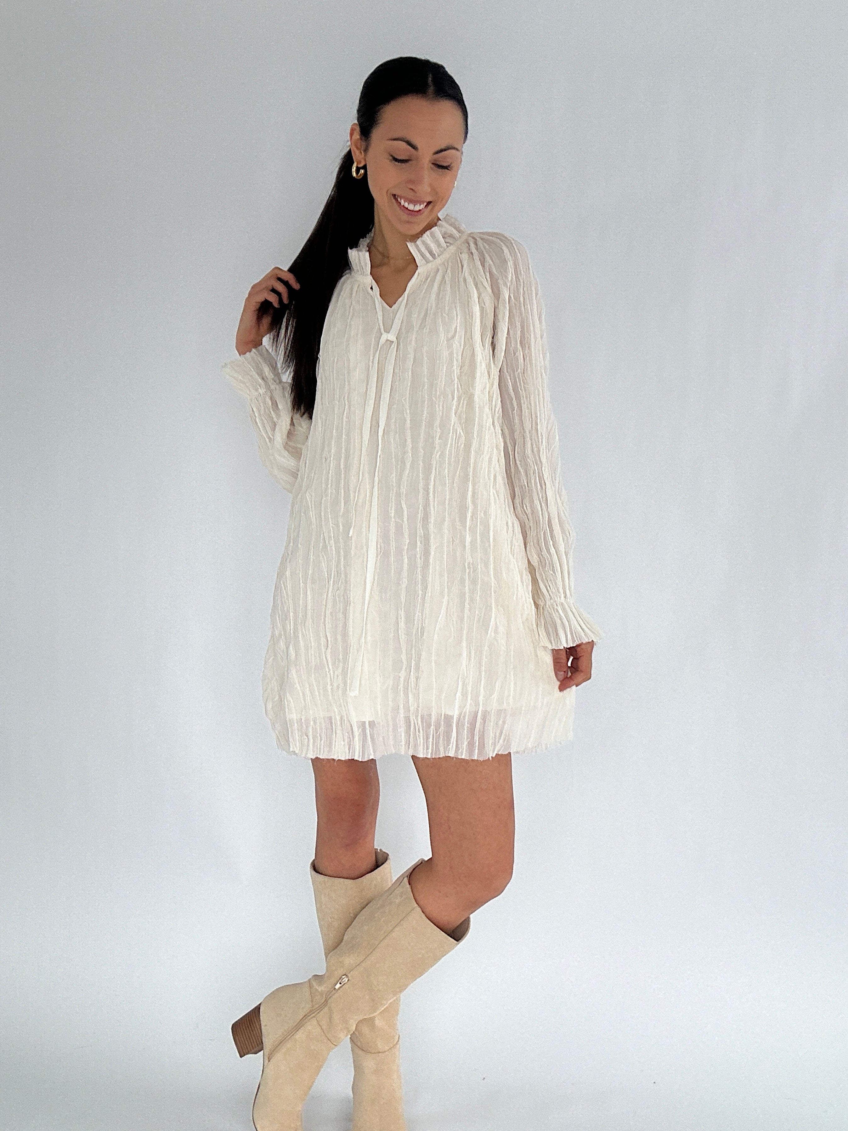 Woman wearing a white pleated dress and beige knee-high boots on a plain background