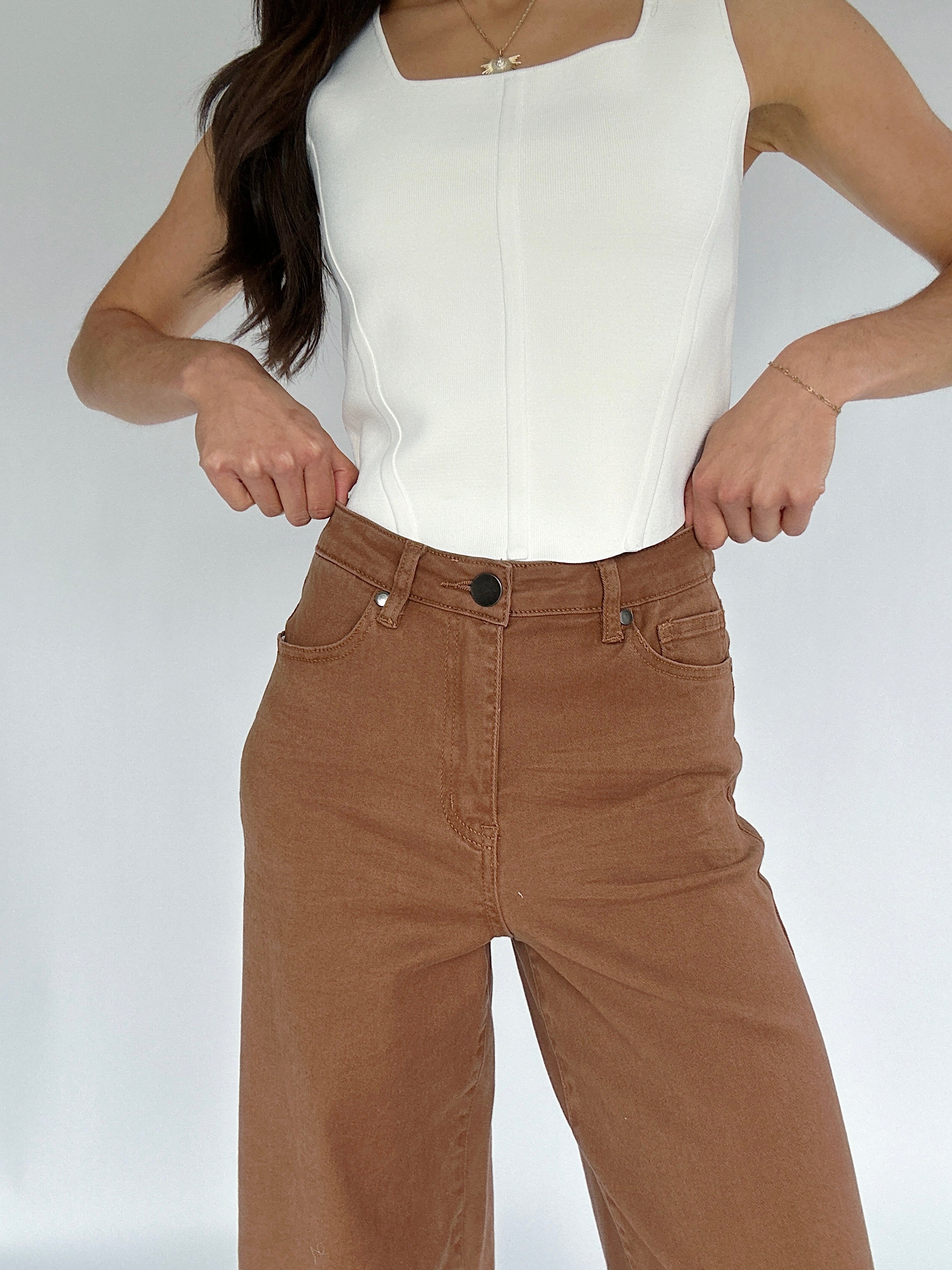 Person wearing a white top and brown pants on a plain background showing zip and button fly