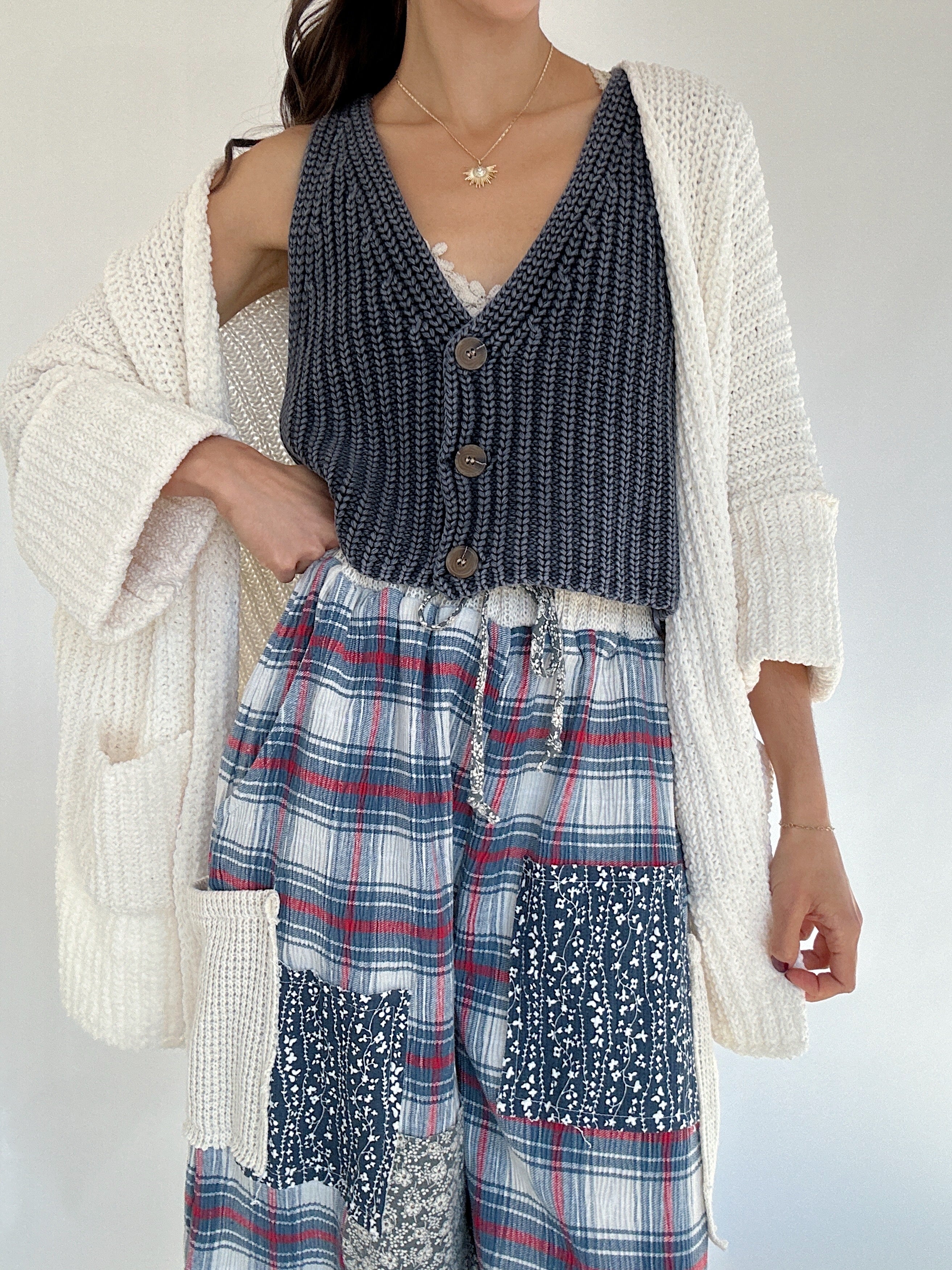 Person wearing a blue knit vest top with button front, plaid pants, and a white cardigan on a plain background