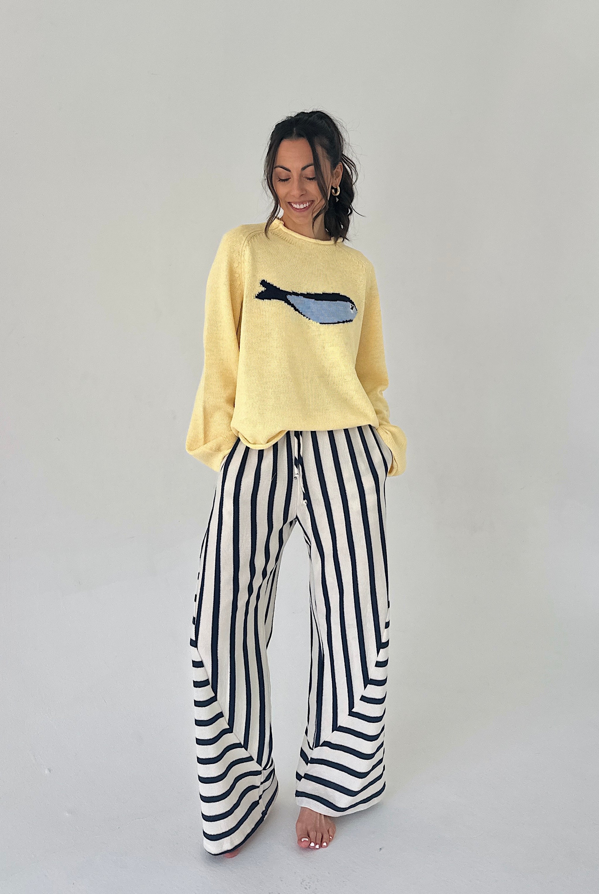 Woman wearing a yellow sweater with a fish design and black and white striped pants on a plain background