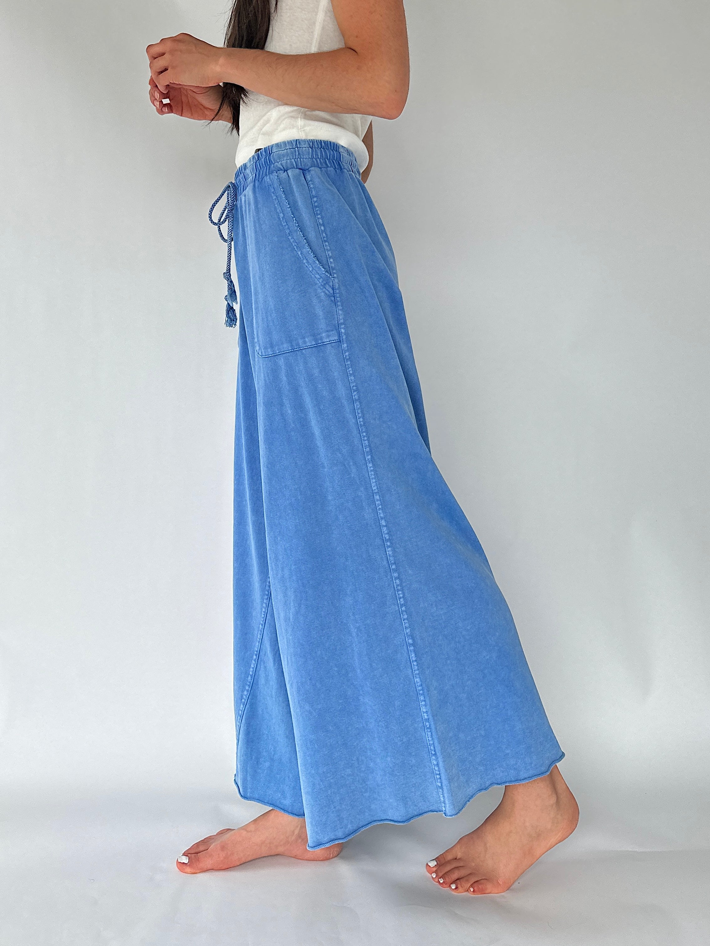 A person wearing a wide-leg pants with a drawstring waist and side pocket, paired with a white sleeveless top, standing barefoot against a plain light gray studio background.