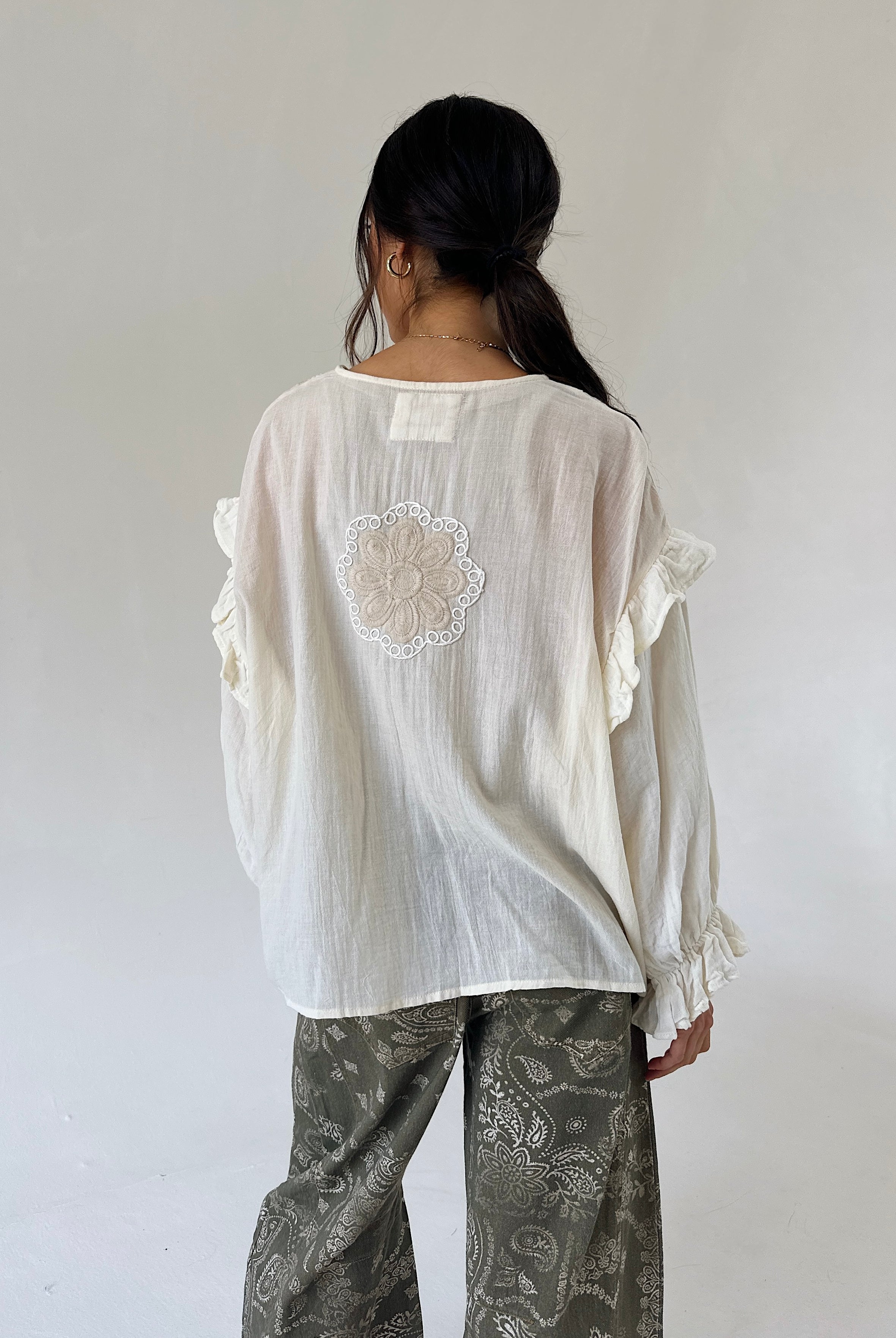 A woman viewed from behind wearing a loose white blouse with ruffled sleeves and a floral lace applique on the back, paired with gray paisley pants, against a plain light gray studio background.