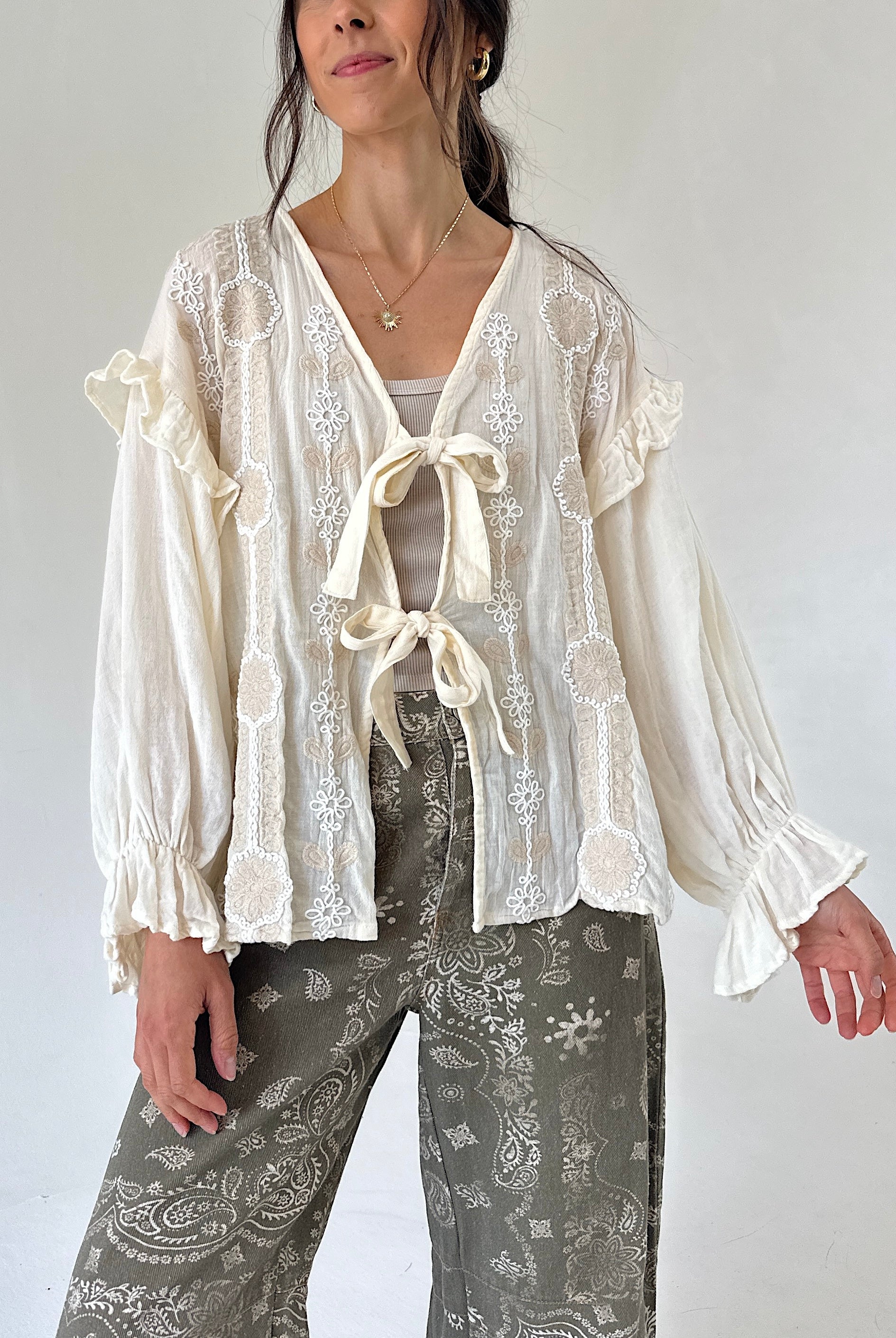 A woman wearing a cream-colored long-sleeve blouse with ruffled cuffs, lace and a front tie, paired with gray paisley pants, against a plain light background.