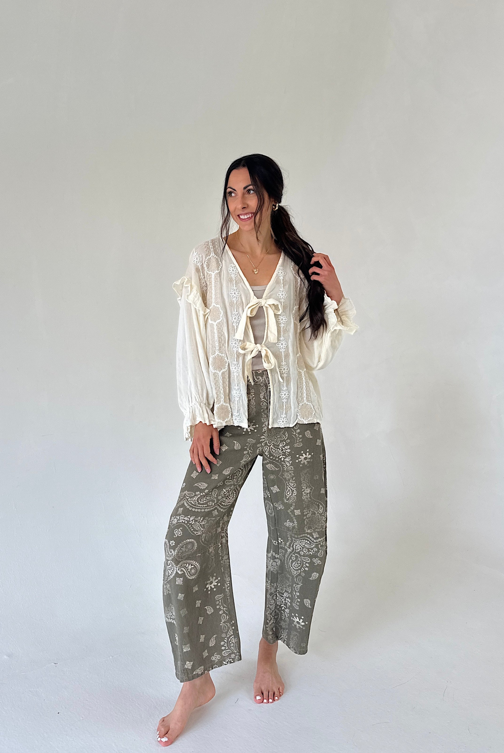 A woman modeling a bohemian-style outfit of a cream ruffled long-sleeve top and olive green patterned wide-leg pants, standing against a plain light gray studio background.